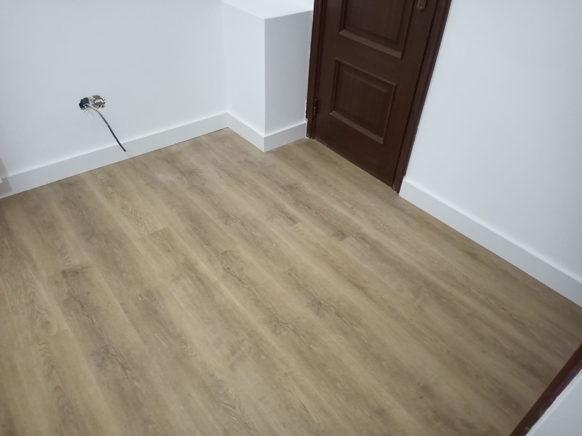 Piso com aparência de madeira em um cômodo com paredes brancas, rodapés e porta marrom.