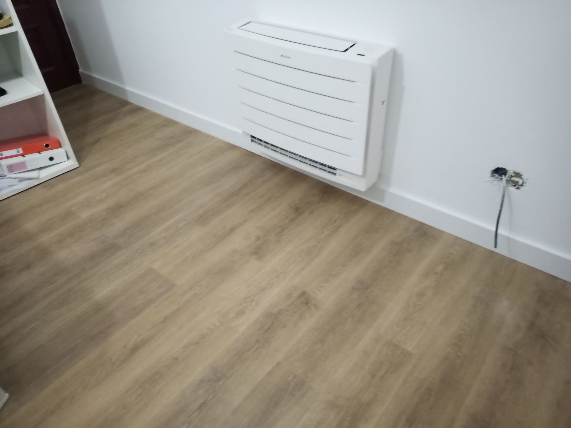 Aquecedor branco de parede acima do piso de madeira. Tomada elétrica visível.