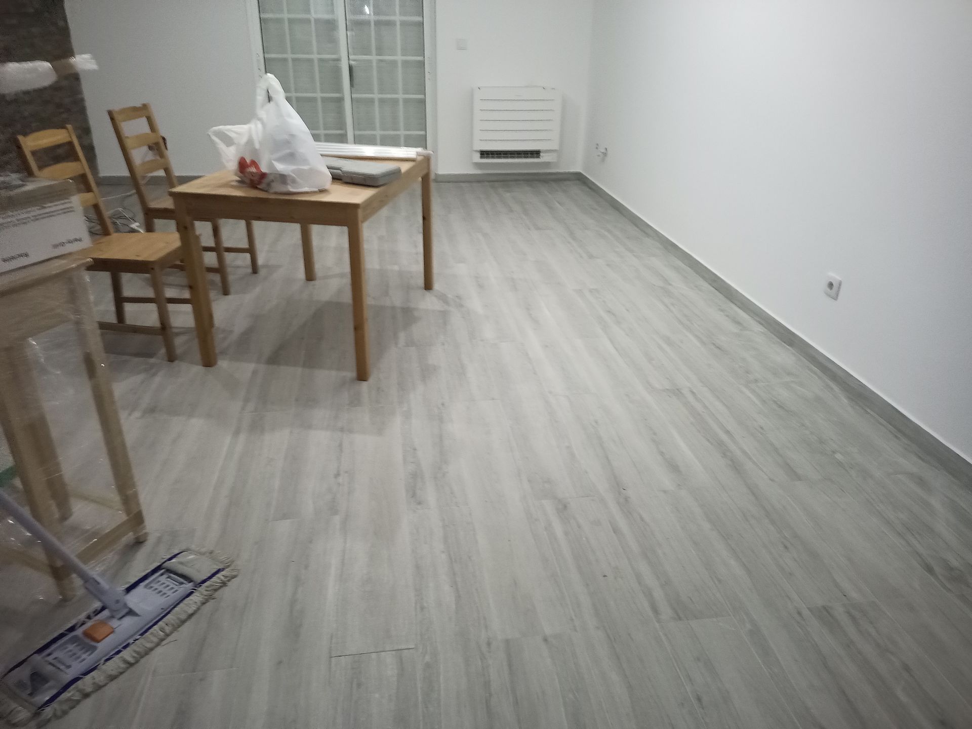 Piso imitando madeira em tom cinza claro em uma sala com mesa, cadeiras e uma parede branca com radiador.