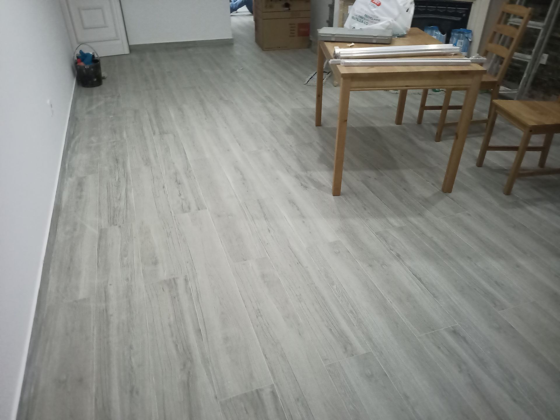 O piso, que imita madeira em tom cinza claro, se estende por toda a sala. Uma mesa e uma cadeira de madeira estão dispostas no chão.