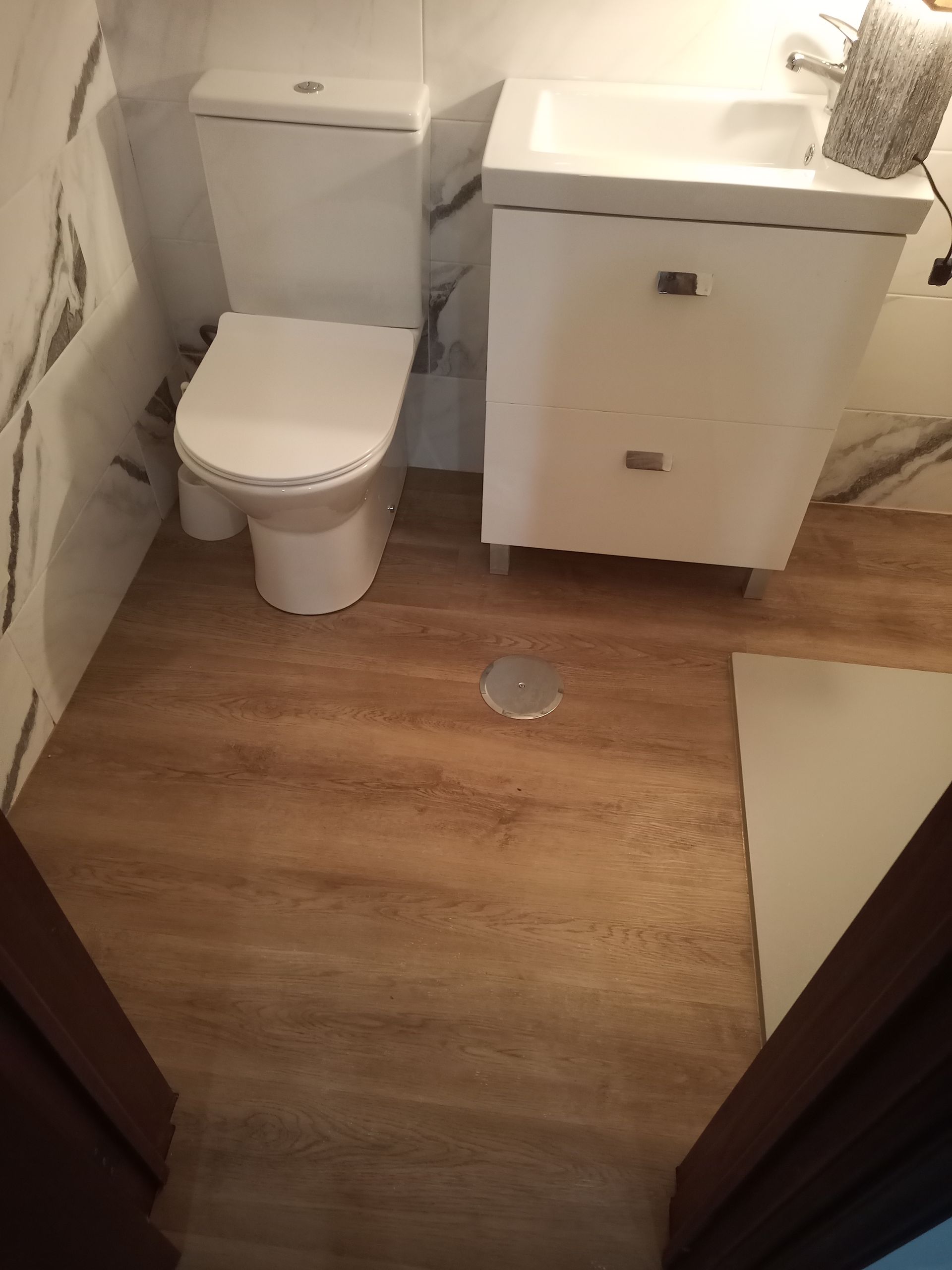 Banheiro com vaso sanitário branco, armário com pia e piso imitando madeira. Parede com aparência de mármore, porta-toalhas.