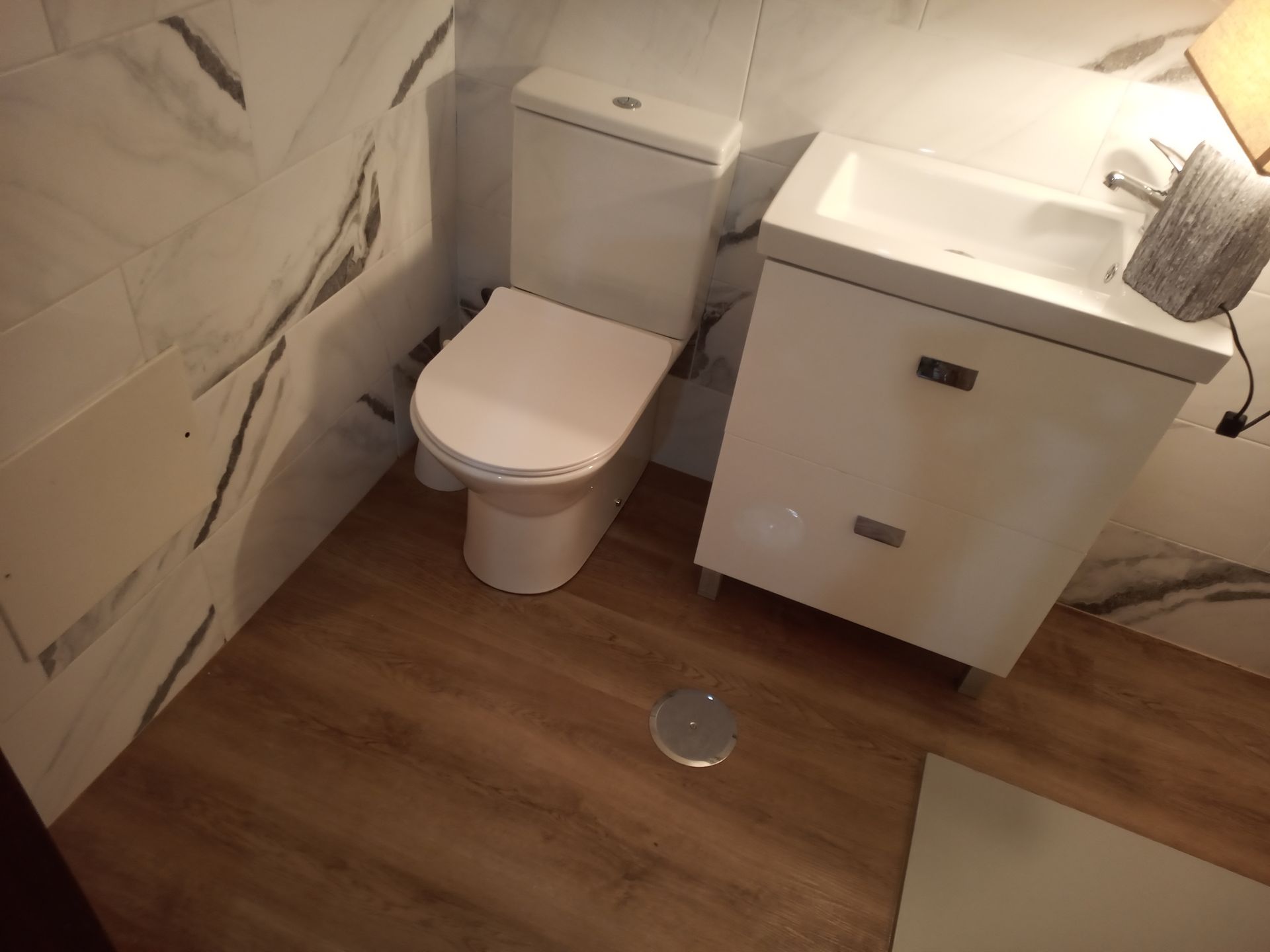 Um banheiro pequeno com vaso sanitário branco, armário com pia e piso que imita madeira marrom. Azulejos brancos que imitam mármore nas paredes.