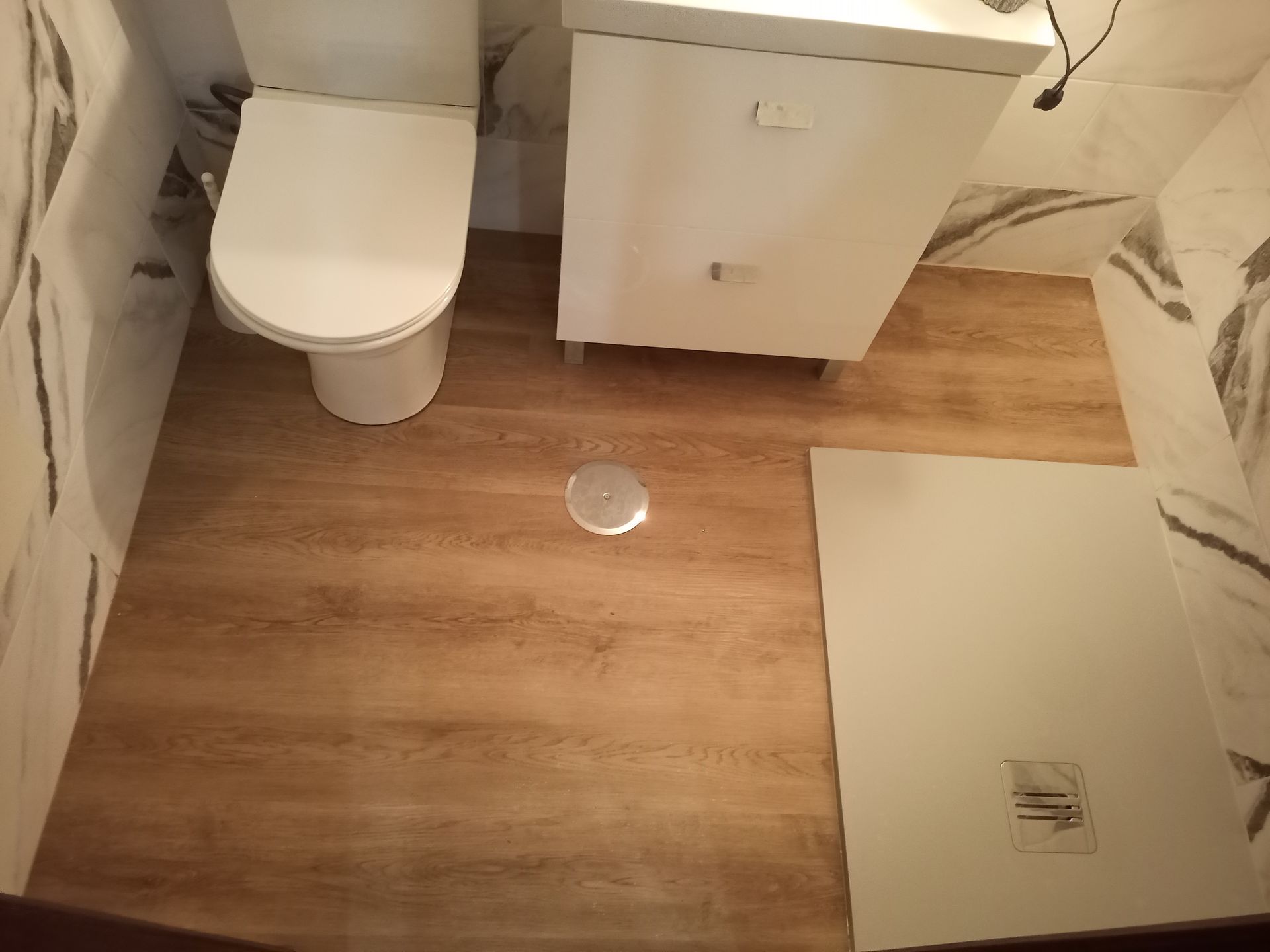 Banheiro com piso imitando madeira, vaso sanitário branco, pia e base de chuveiro. Paredes com estampa de mármore.