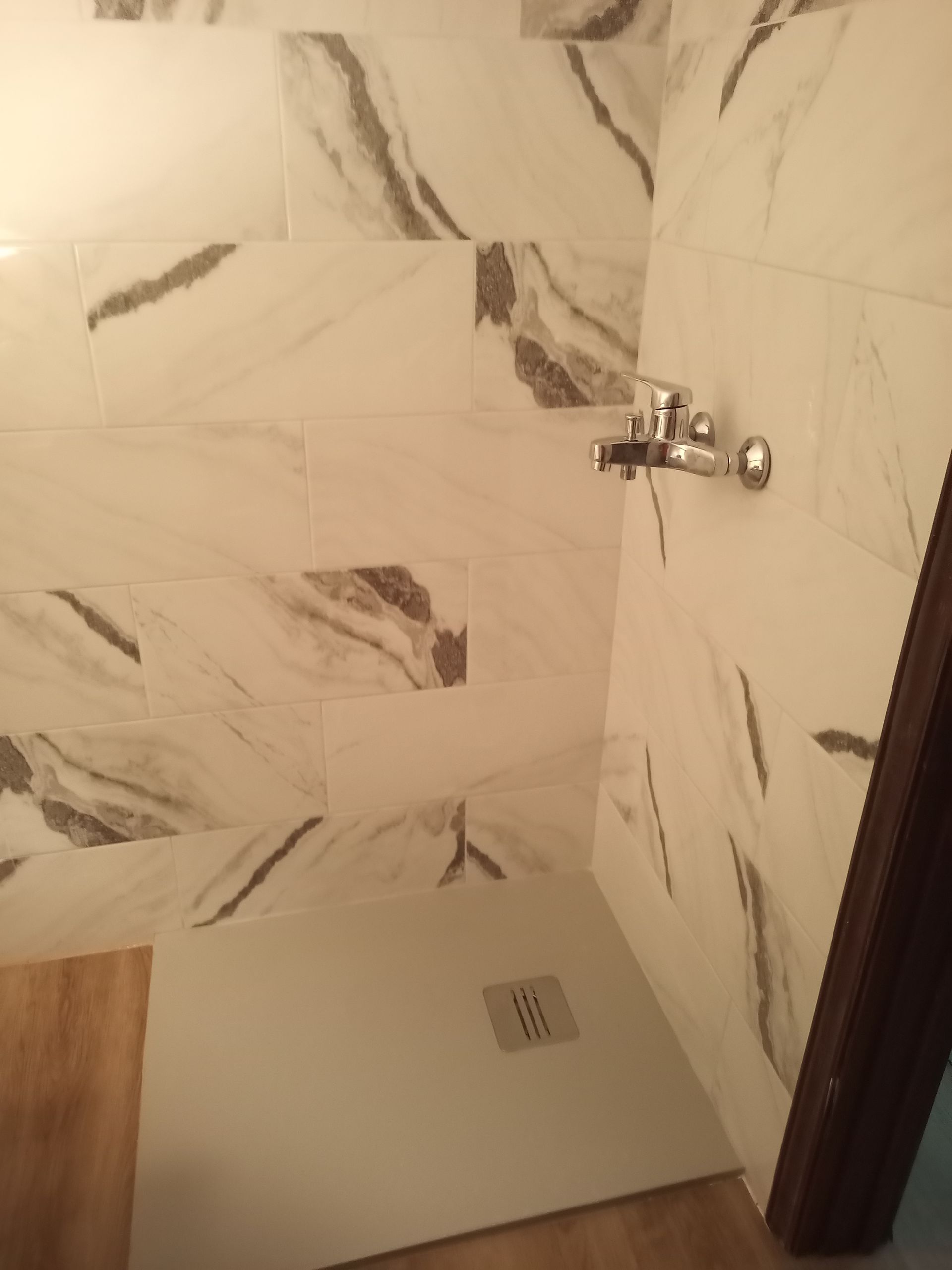 Cabine de duche com azulejos brancos com padrão de mármore e acessórios cromados. A base do chuveiro está visível.