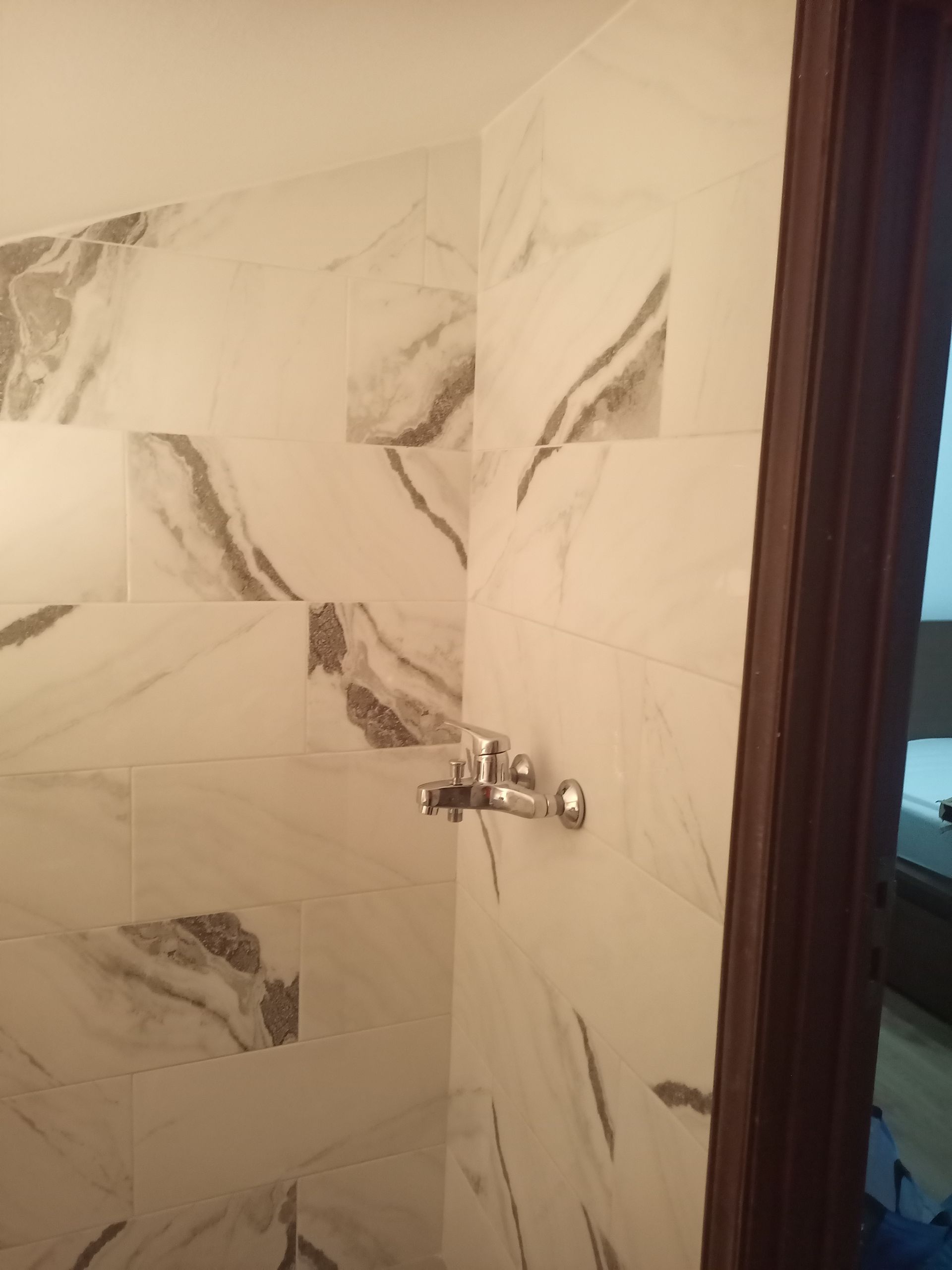 Chuveiro revestido com azulejos de mármore branco e acessórios prateados, vista de canto. Batente da porta marrom à direita.