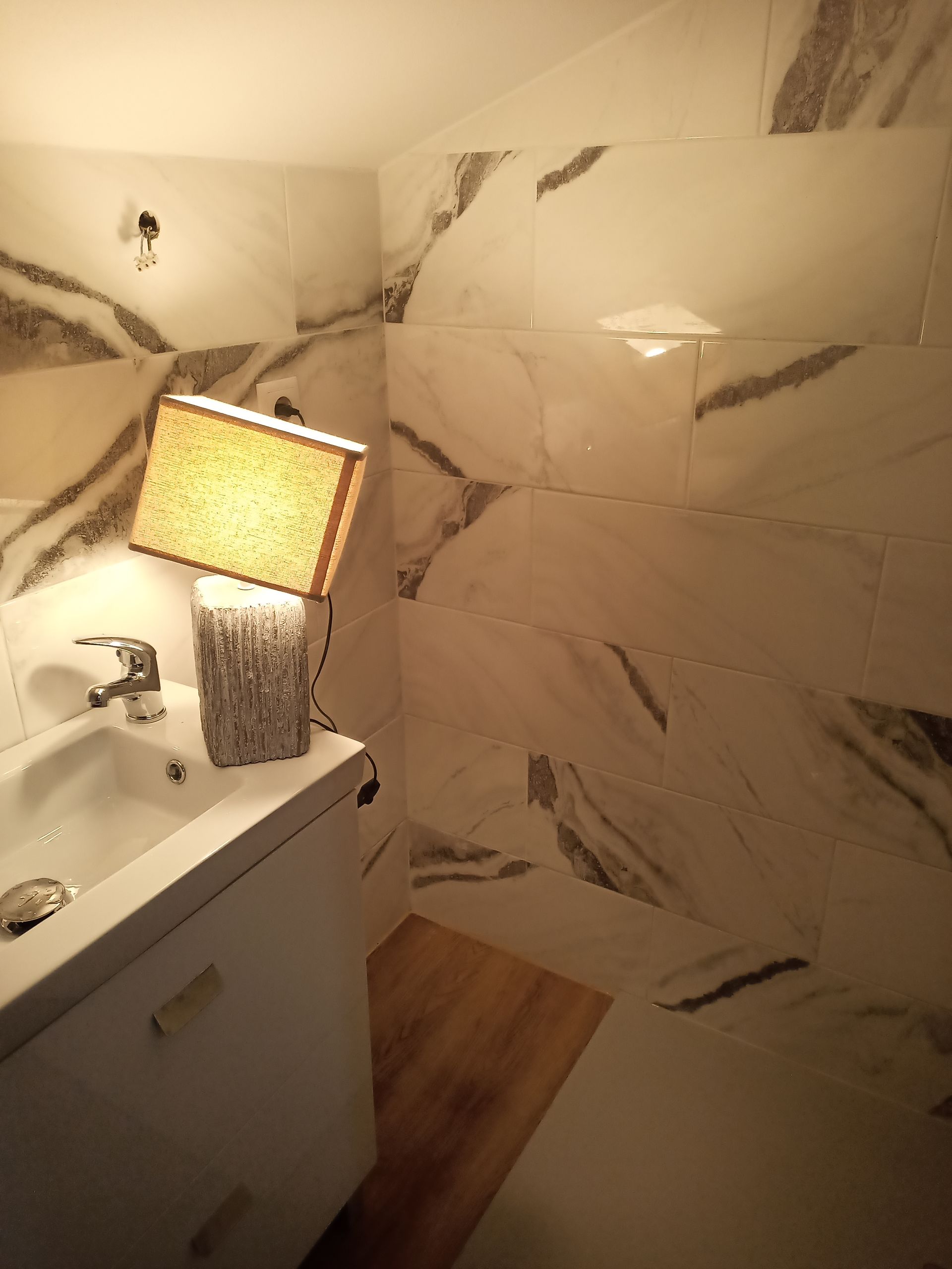 Banheiro com paredes revestidas com azulejos de mármore branco, pia branca e luminária sobre a bancada.