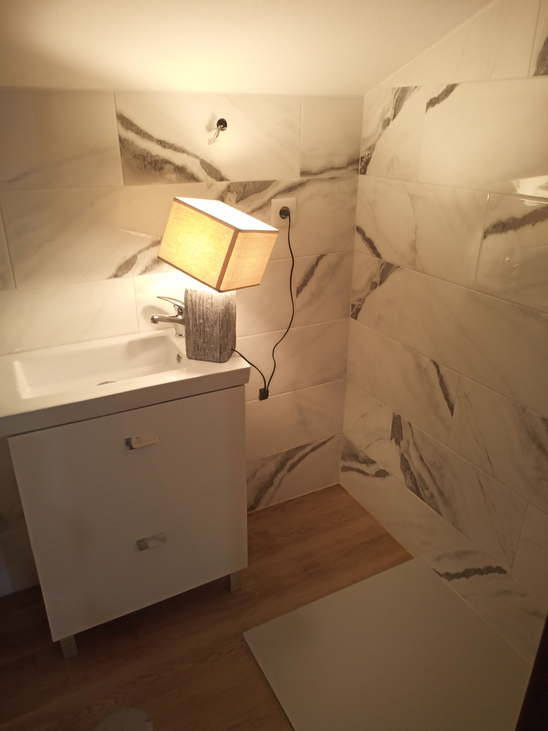 Banheiro com pia branca, luminária e azulejos que imitam mármore.