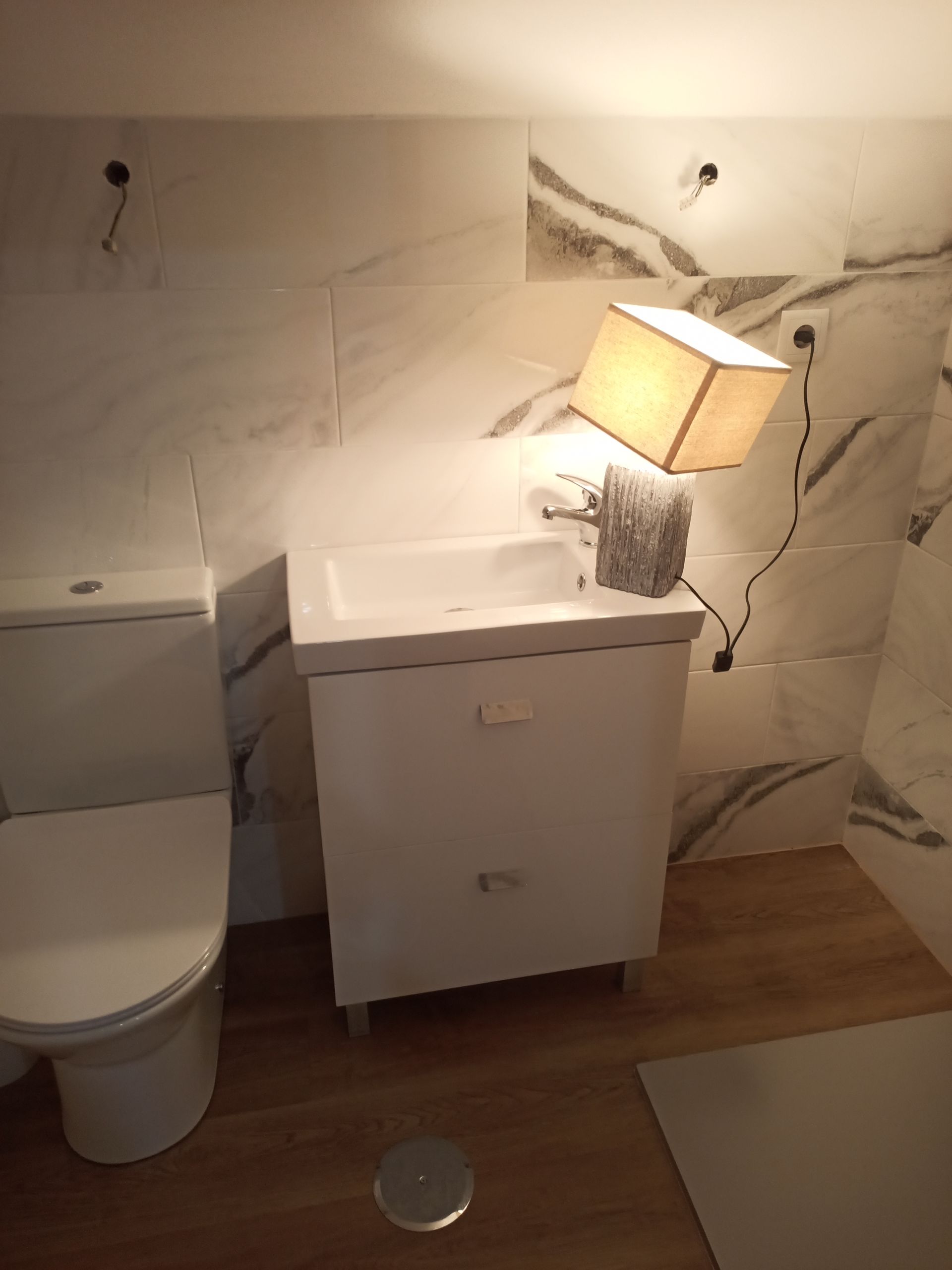 Banheiro com vaso sanitário branco, armário com pia e azulejos com aparência de mármore. Uma luminária está posicionada precariamente acima da pia.