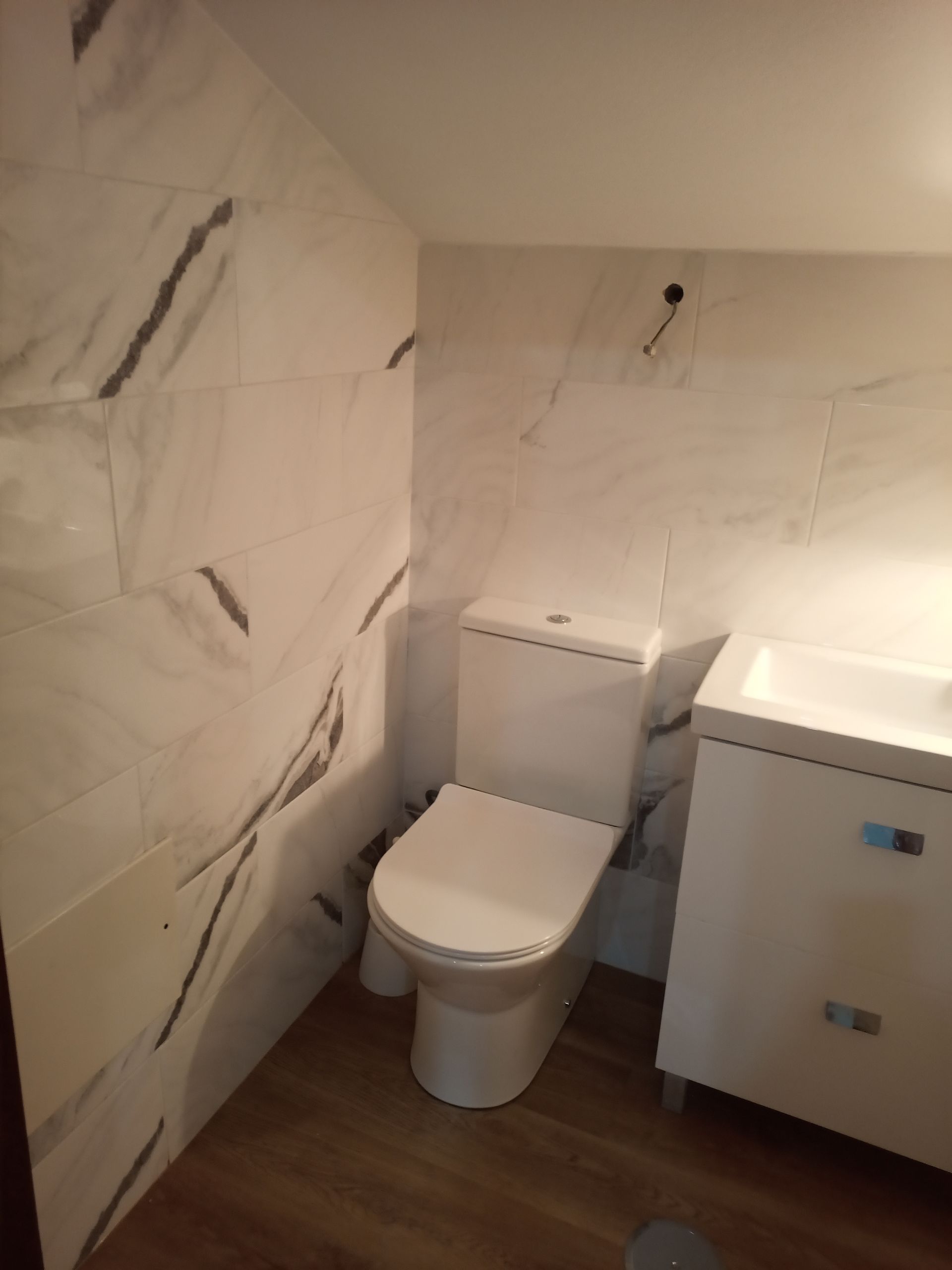 Banheiro com paredes revestidas com azulejos brancos imitando mármore, vaso sanitário branco e pia com armário branco. Piso de madeira.