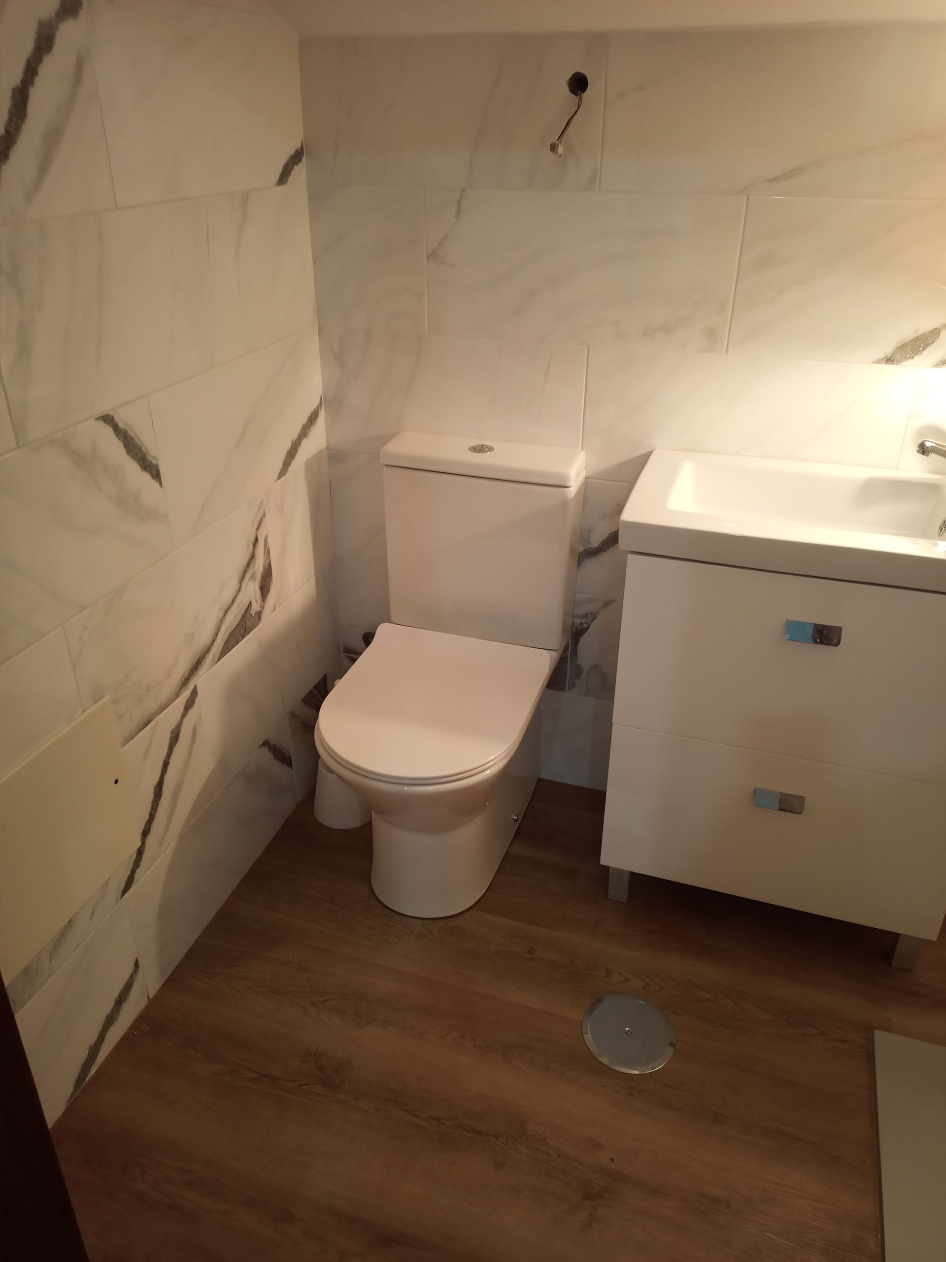 Banheiro com paredes revestidas com azulejos brancos imitando mármore, piso de madeira, vaso sanitário e pia branca.