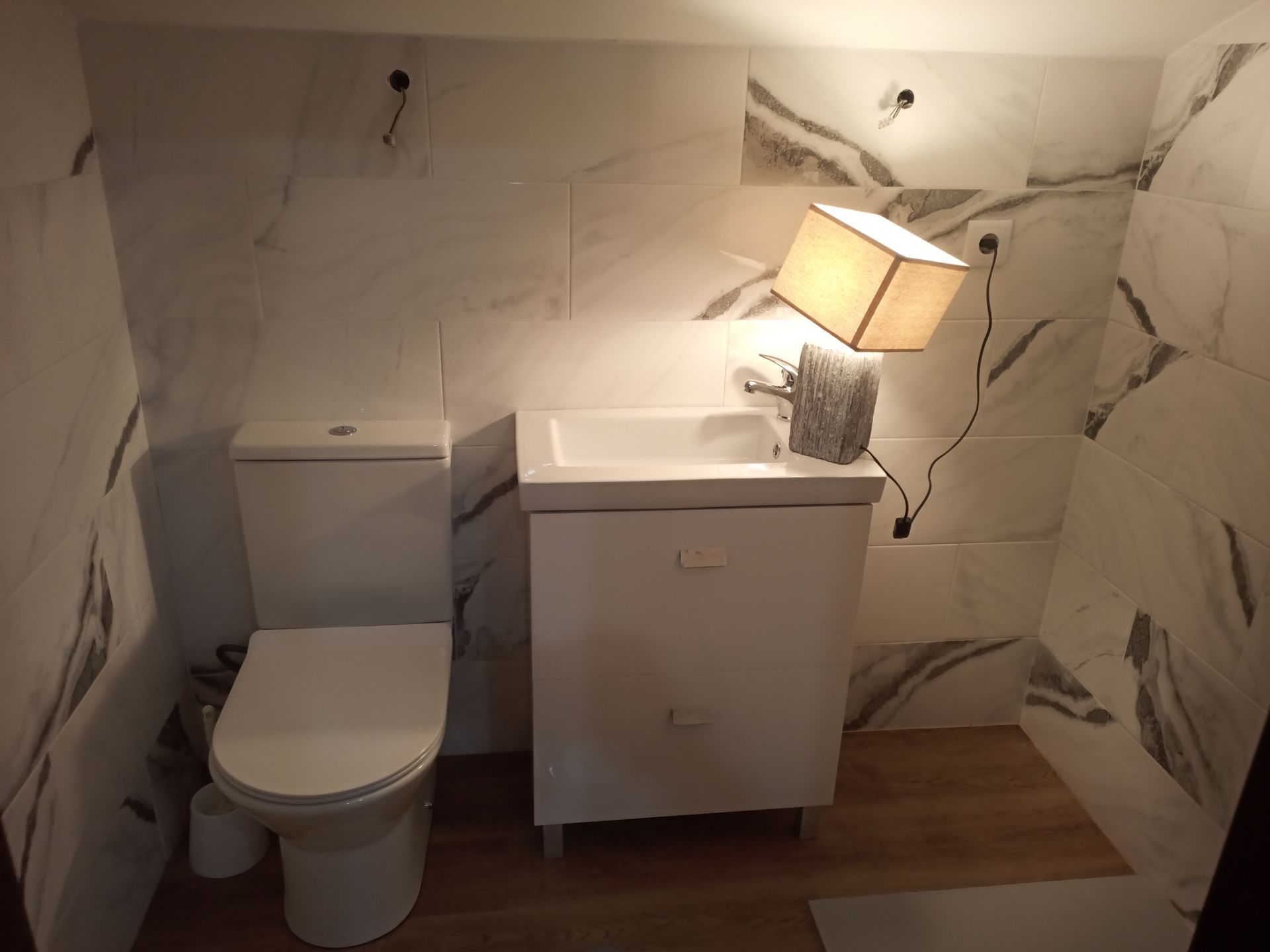 Pequeno banheiro branco com vaso sanitário, pia e luminária em frente a paredes revestidas com azulejos que imitam mármore.