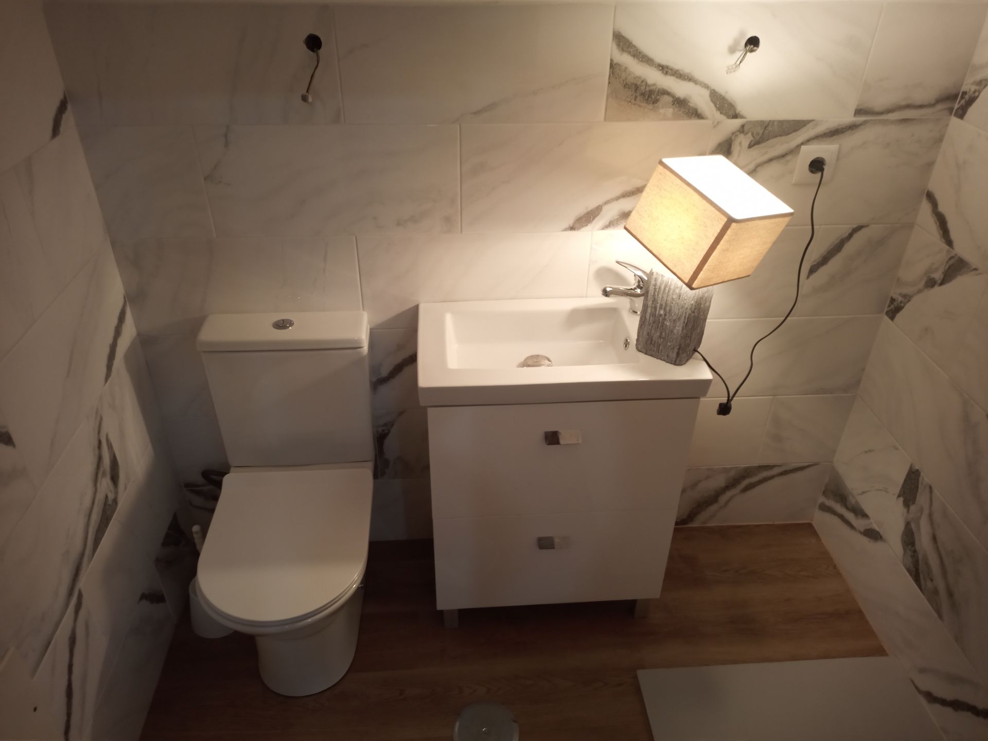 Banheiro pequeno com vaso sanitário branco, pia e azulejos decorativos com estampa de mármore. Uma luminária está sobre a pia.