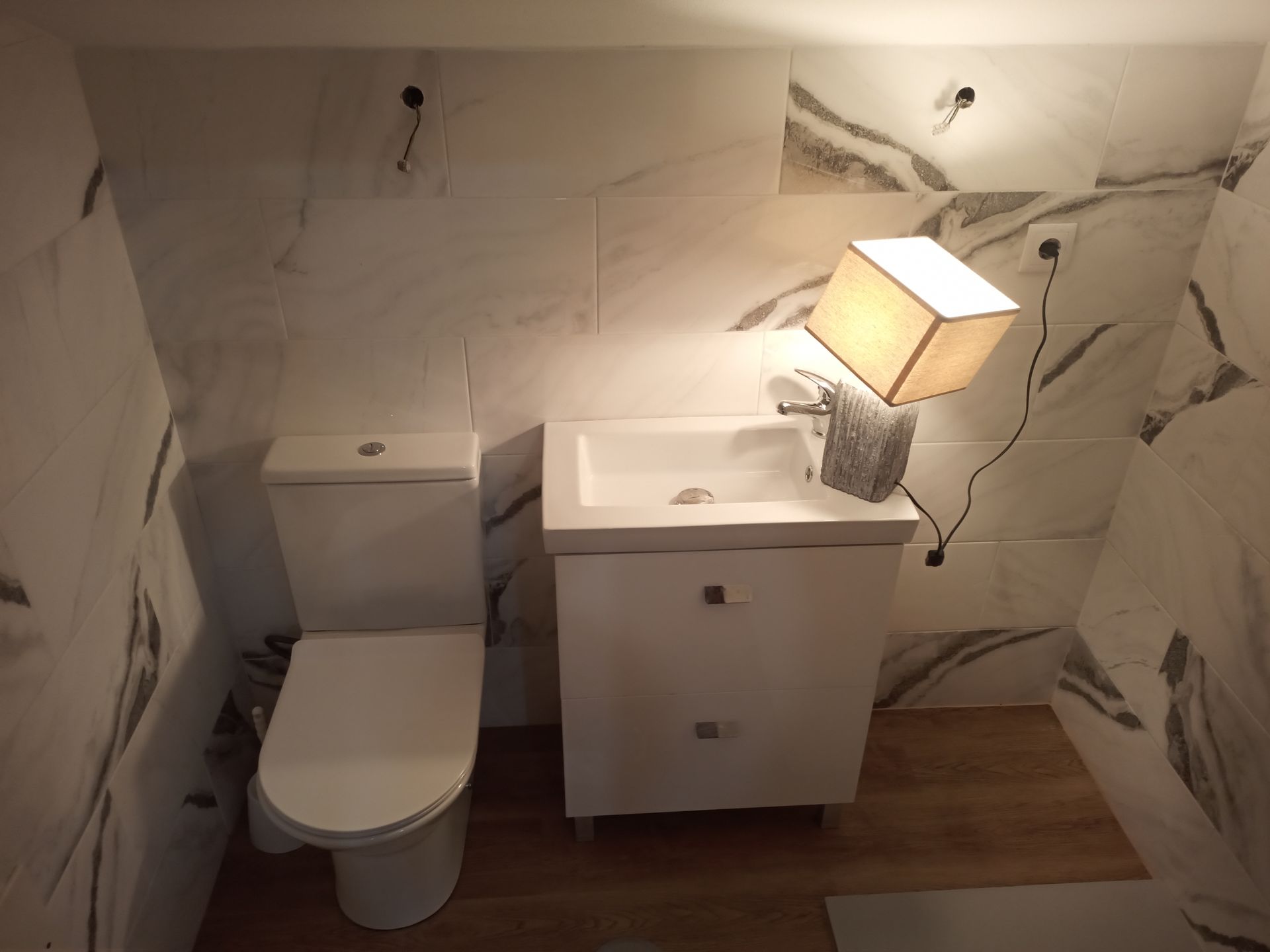 Banheiro pequeno com vaso sanitário branco, pia, azulejos decorativos com estampa de mármore, piso de madeira e um abajur sobre a pia.