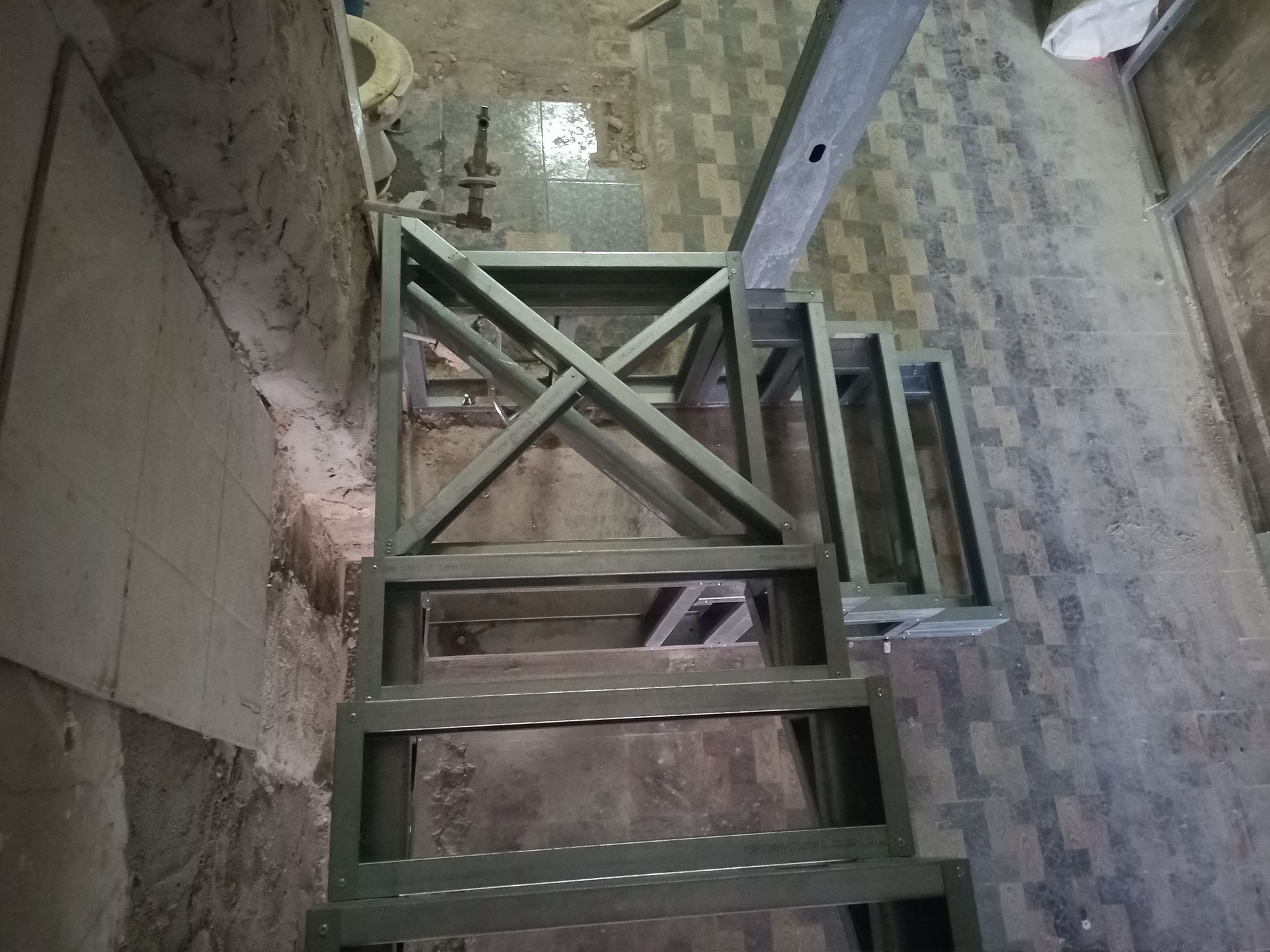 Estrutura metálica de escada em construção, instalada em um edifício com paredes de concreto e piso de azulejo.