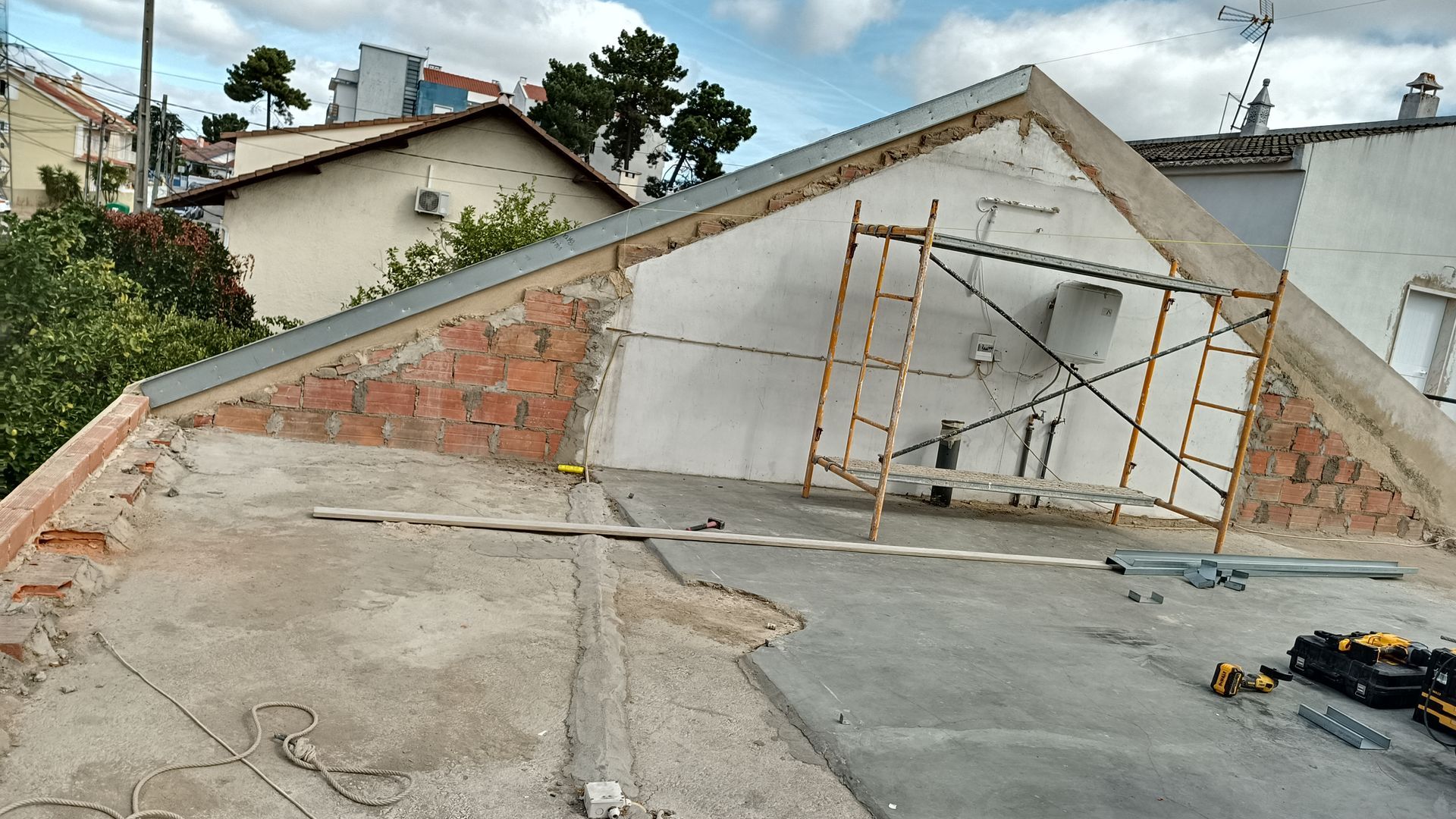 Construção no telhado: Andaimes contra uma parede branca com tijolos aparentes; telhado cinza; edifícios circundantes visíveis.