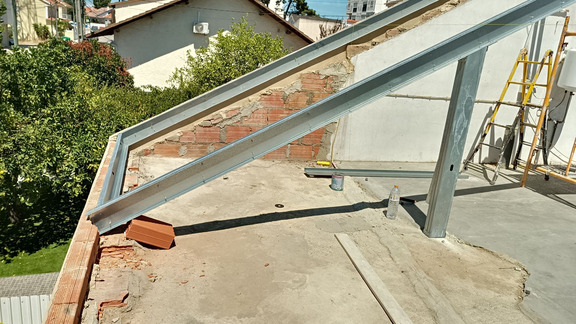 Estrutura metálica sobre um telhado de tijolo e concreto, suportando a instalação de painéis solares, com tijolos e árvores ao fundo.
