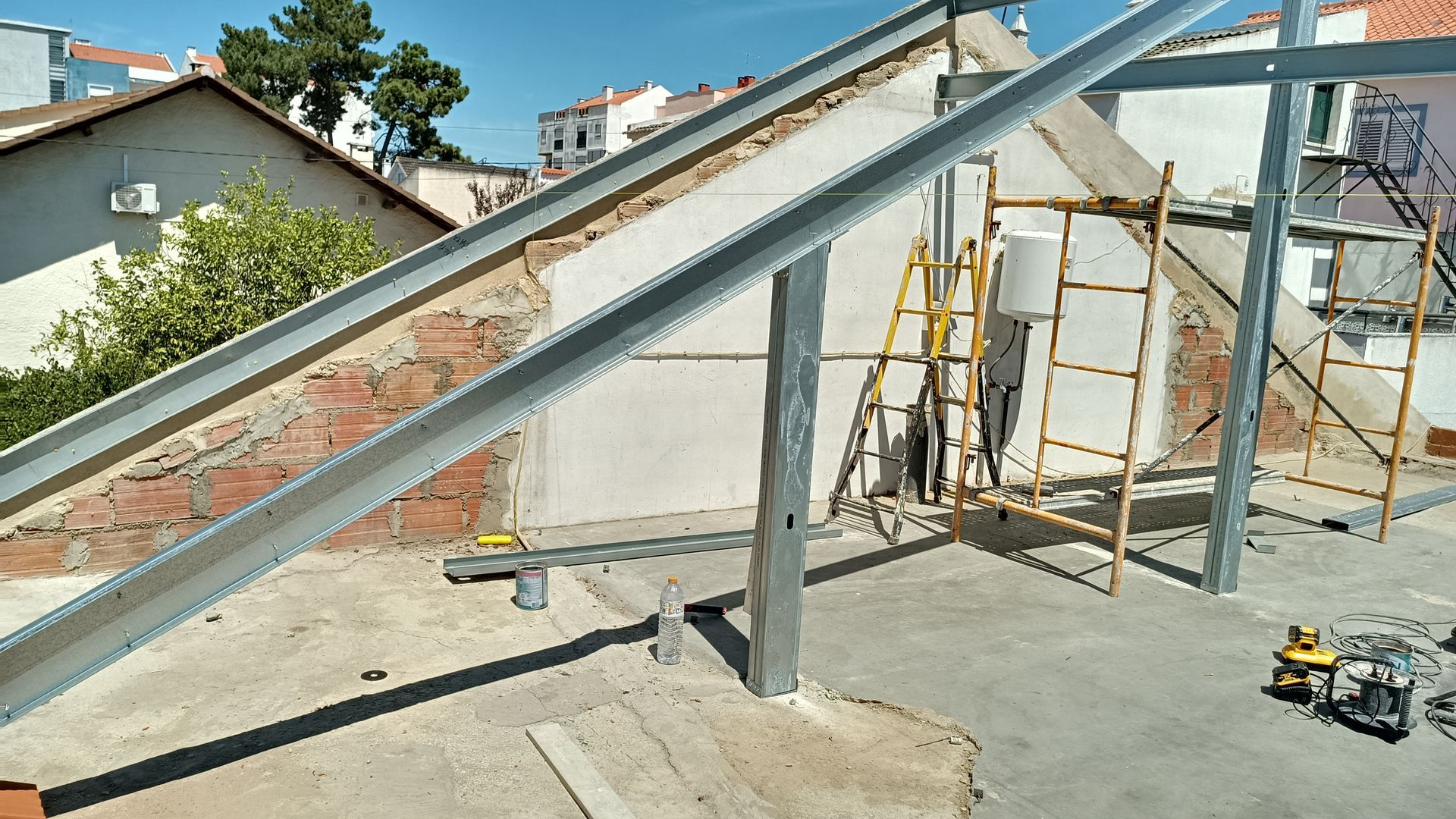 Estrutura de aço sendo erguida em um telhado, com andaimes e uma antiga parede de tijolos ao fundo.