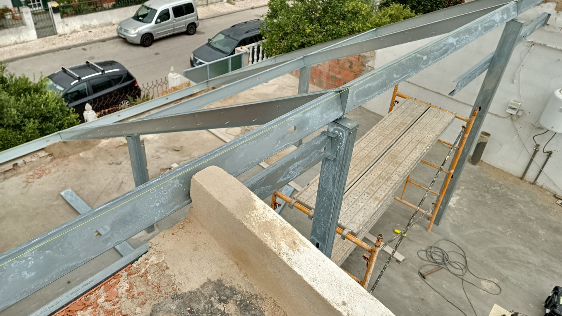 Estrutura metálica em construção no telhado, andaimes, tábuas, carros na rua abaixo.