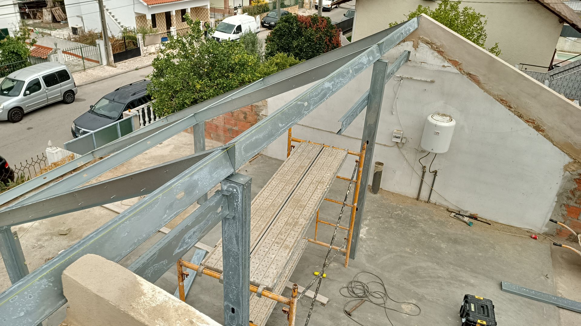 Estrutura metálica de um telhado em construção; andaimes e um aquecedor de água são visíveis.