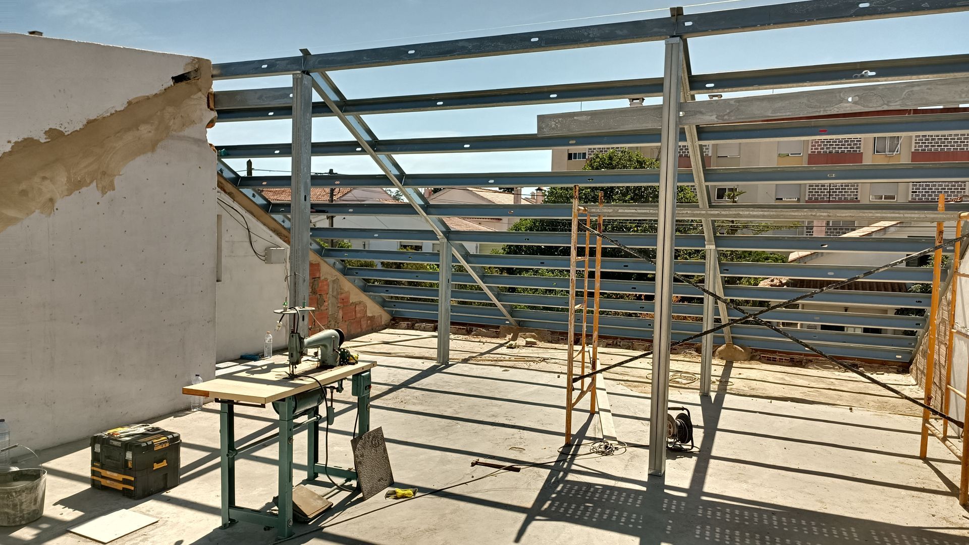 Estrutura metálica em um telhado. Uma máquina de costura está sobre uma mesa. A luz do sol projeta sombras. Prédios visíveis ao fundo.