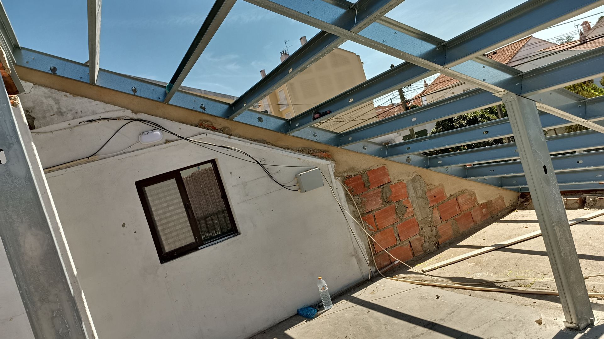 Estrutura de aço sobre um edifício com parede branca, tijolos e uma janela.