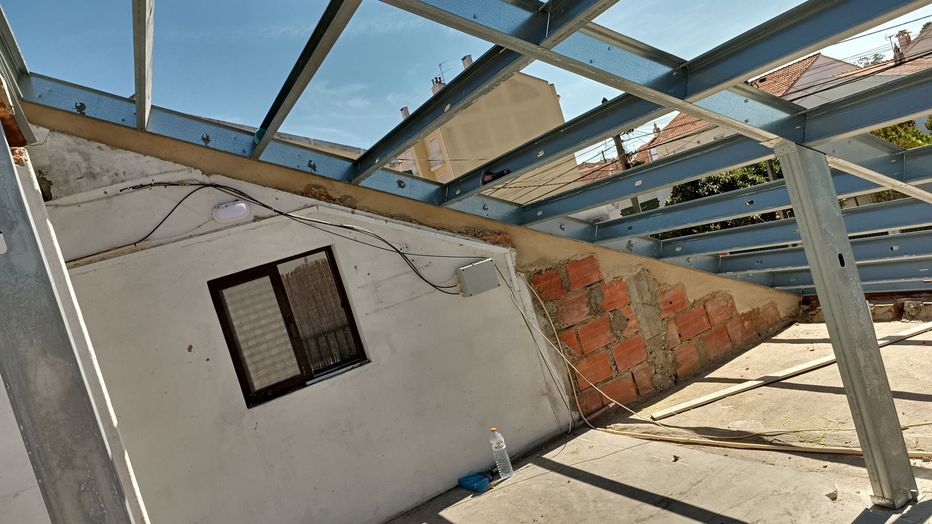 Canteiro de obras: estrutura metálica sobre uma parede de tijolos e branca com uma janela. Céu azul.
