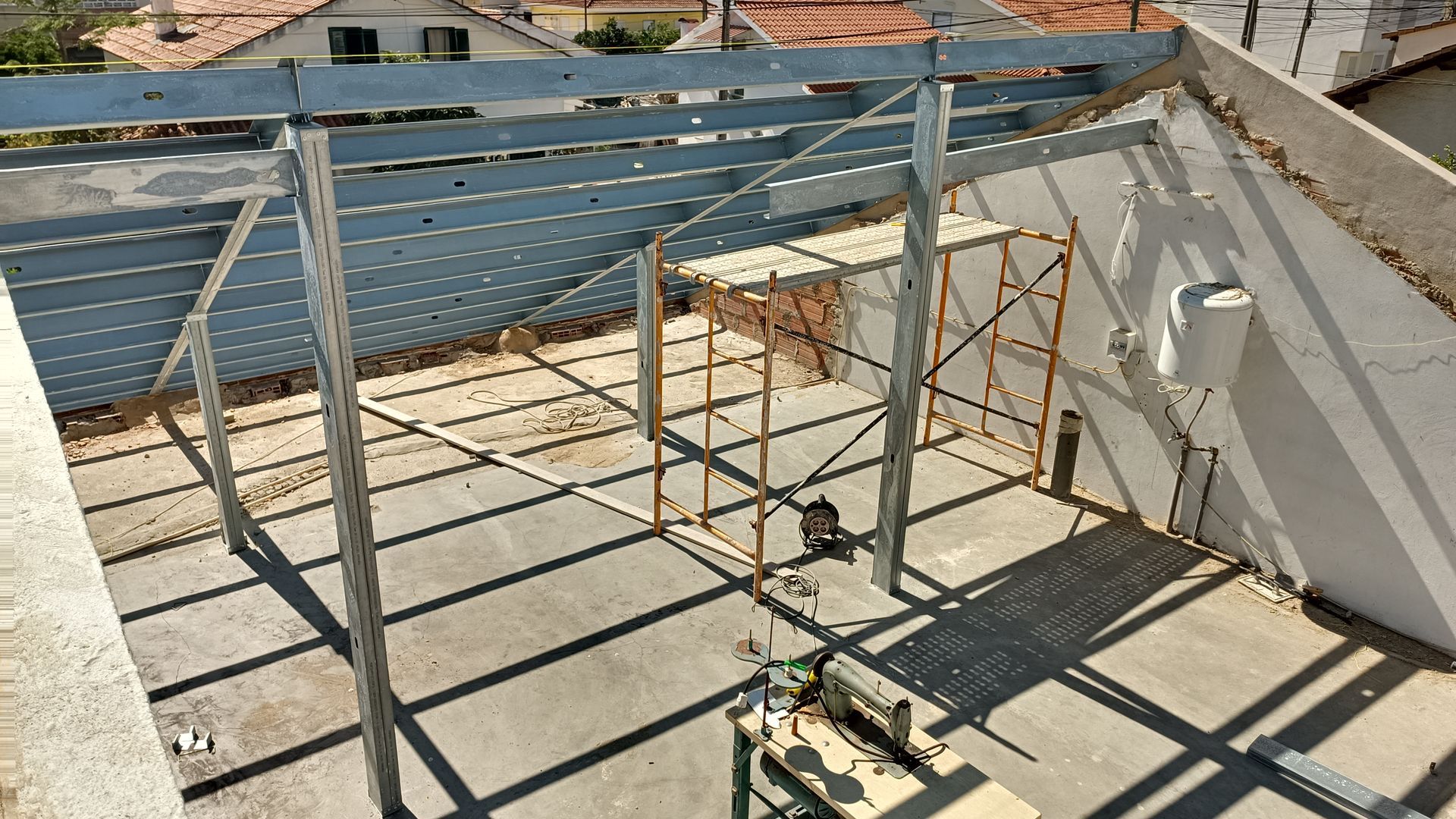Canteiro de obras: estrutura metálica de telhado sendo construída sobre uma cobertura plana, com andaimes no interior.