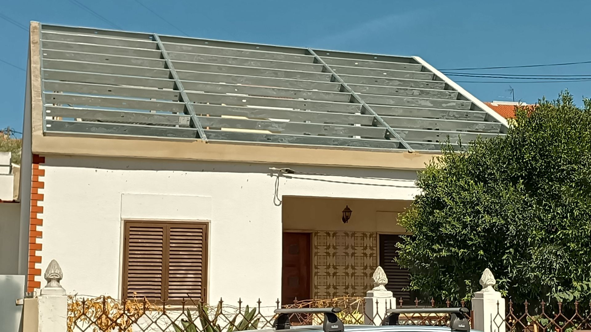 Casa com estrutura metálica no telhado. Exterior branco, persianas marrons, arbusto verde, céu azul.