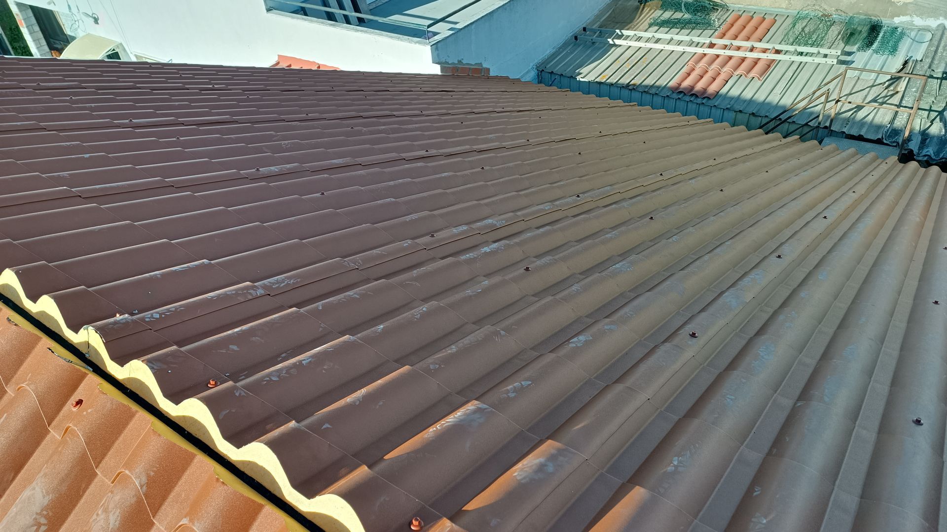 Telhado ondulado, marrom-avermelhado, de um edifício, com um telhado de cor mais clara visível ao fundo.