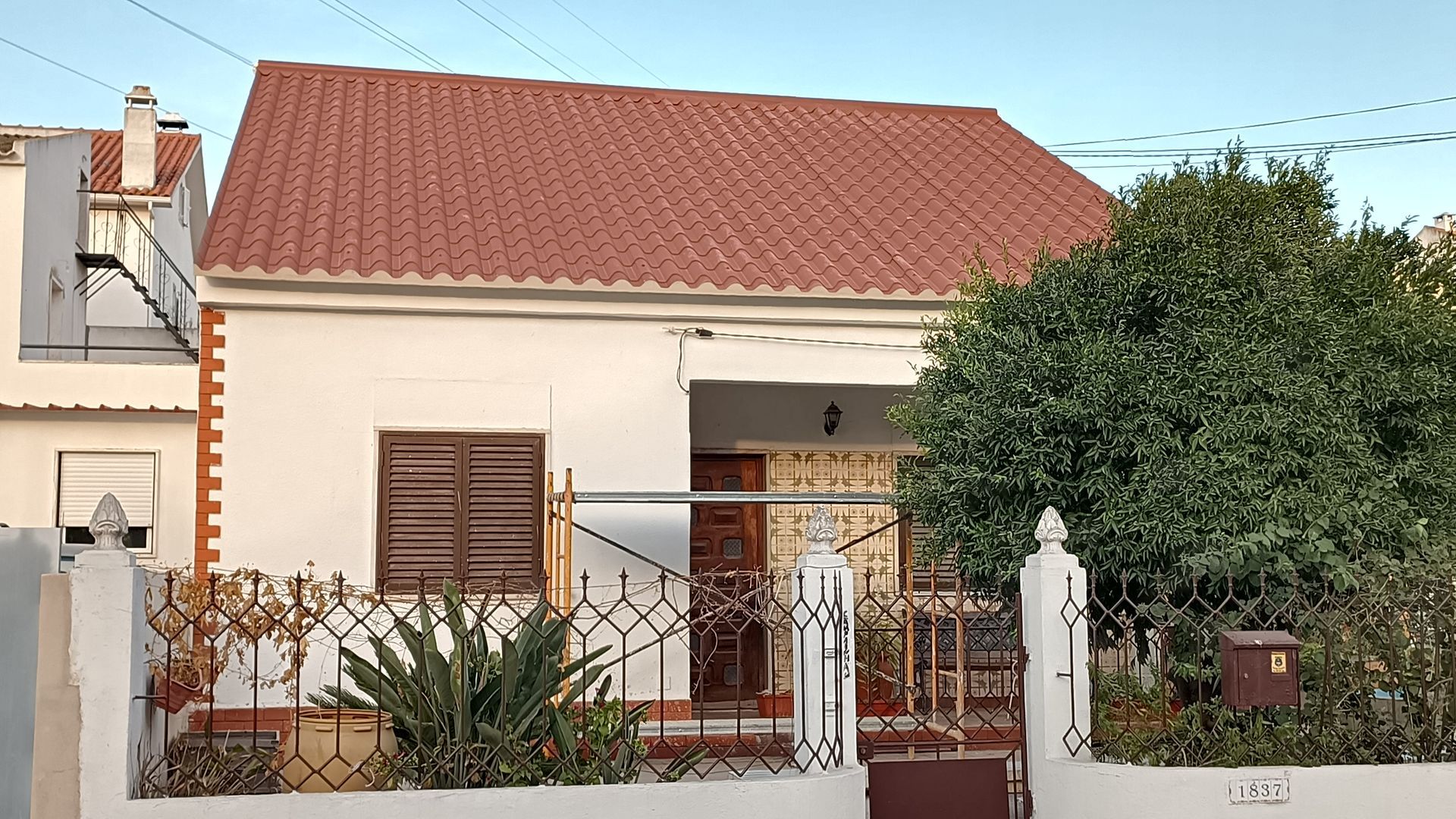 Casa branca com telhado de telha vermelha, venezianas marrons e cerca de ferro forjado.