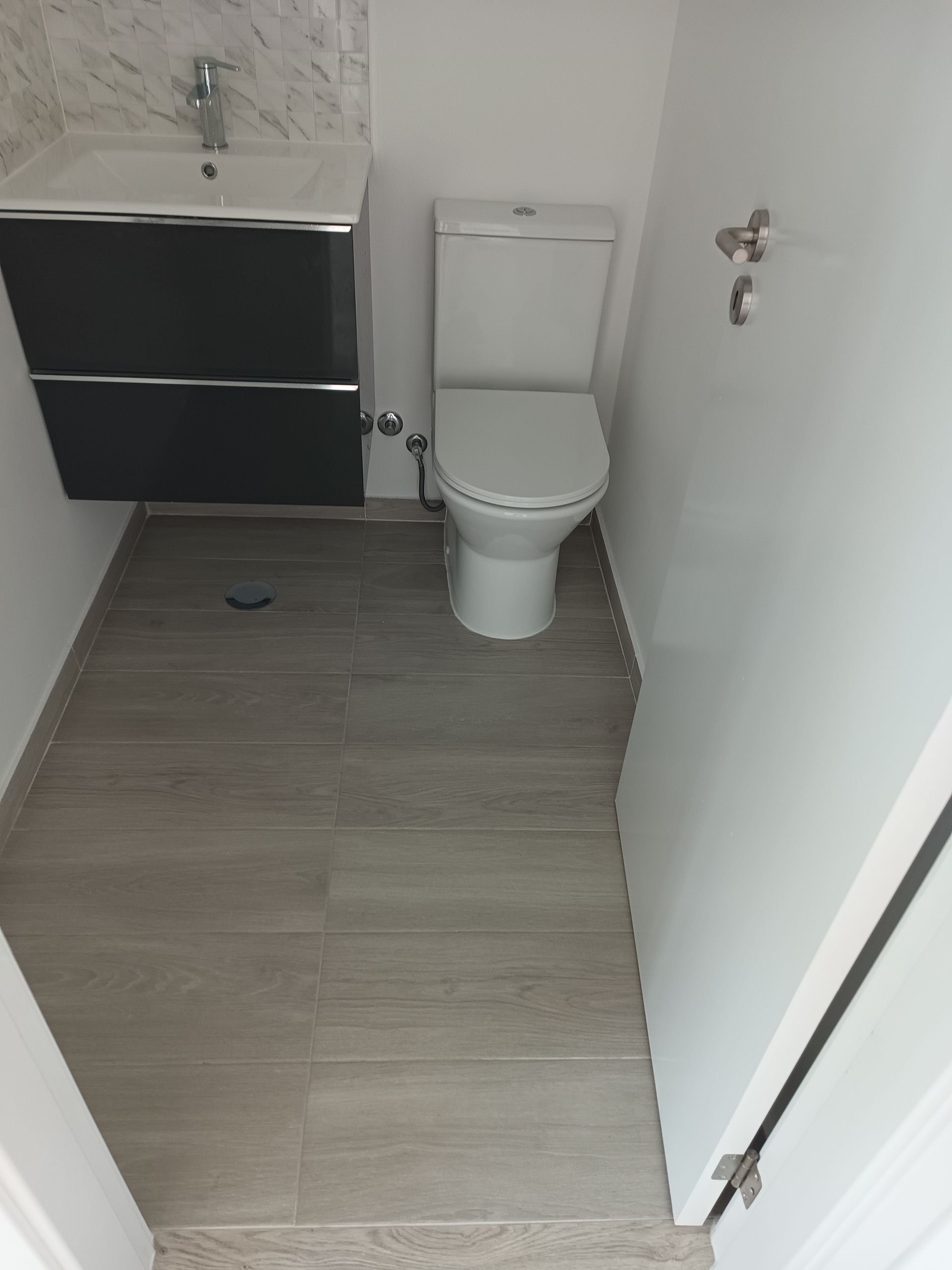 Banheiro pequeno com vaso sanitário e pia. Piso de azulejo imitando madeira cinza e paredes brancas.