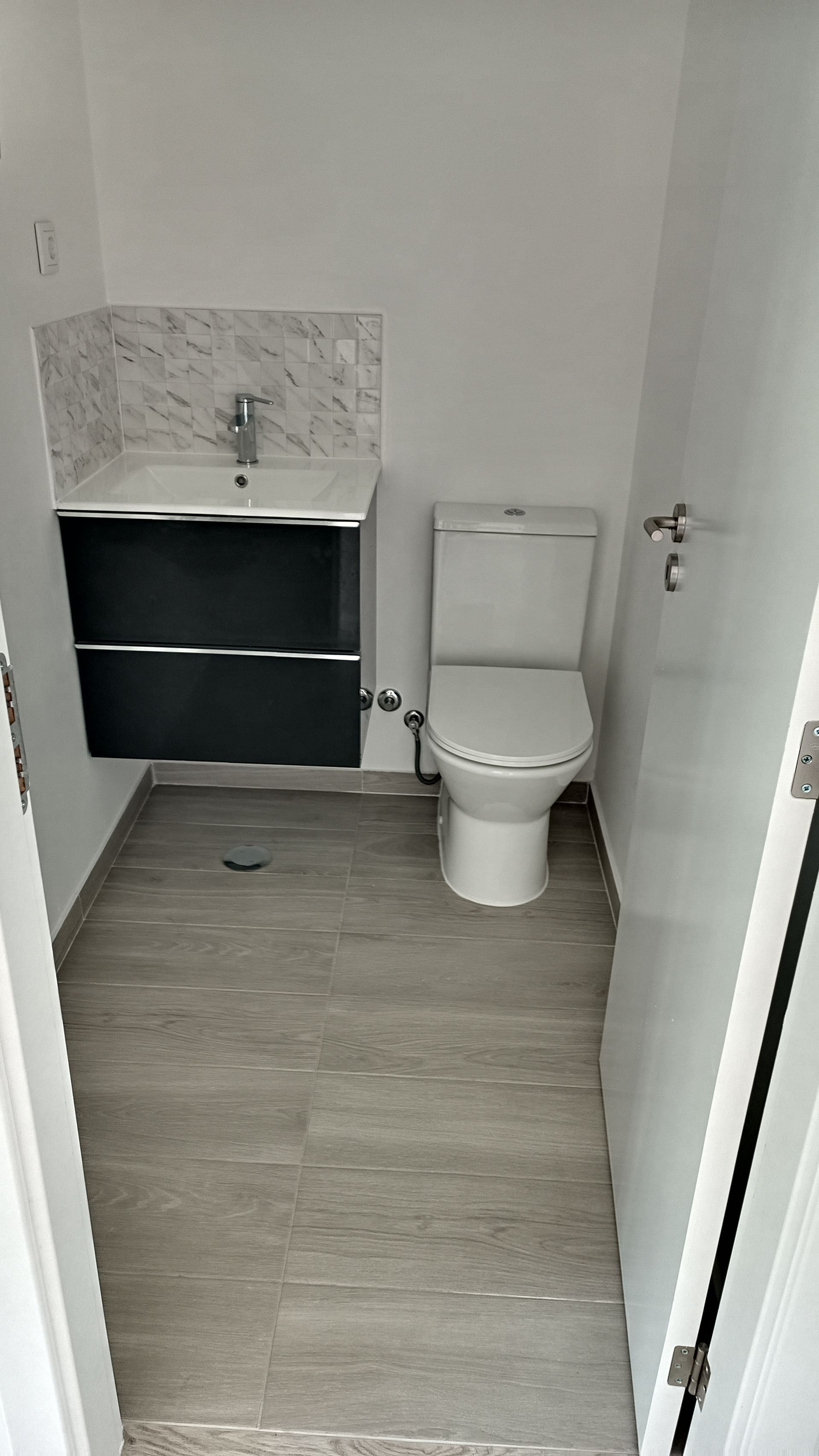 Banheiro pequeno com vaso sanitário e pia. Bancada preta, piso cinza e paredes brancas.