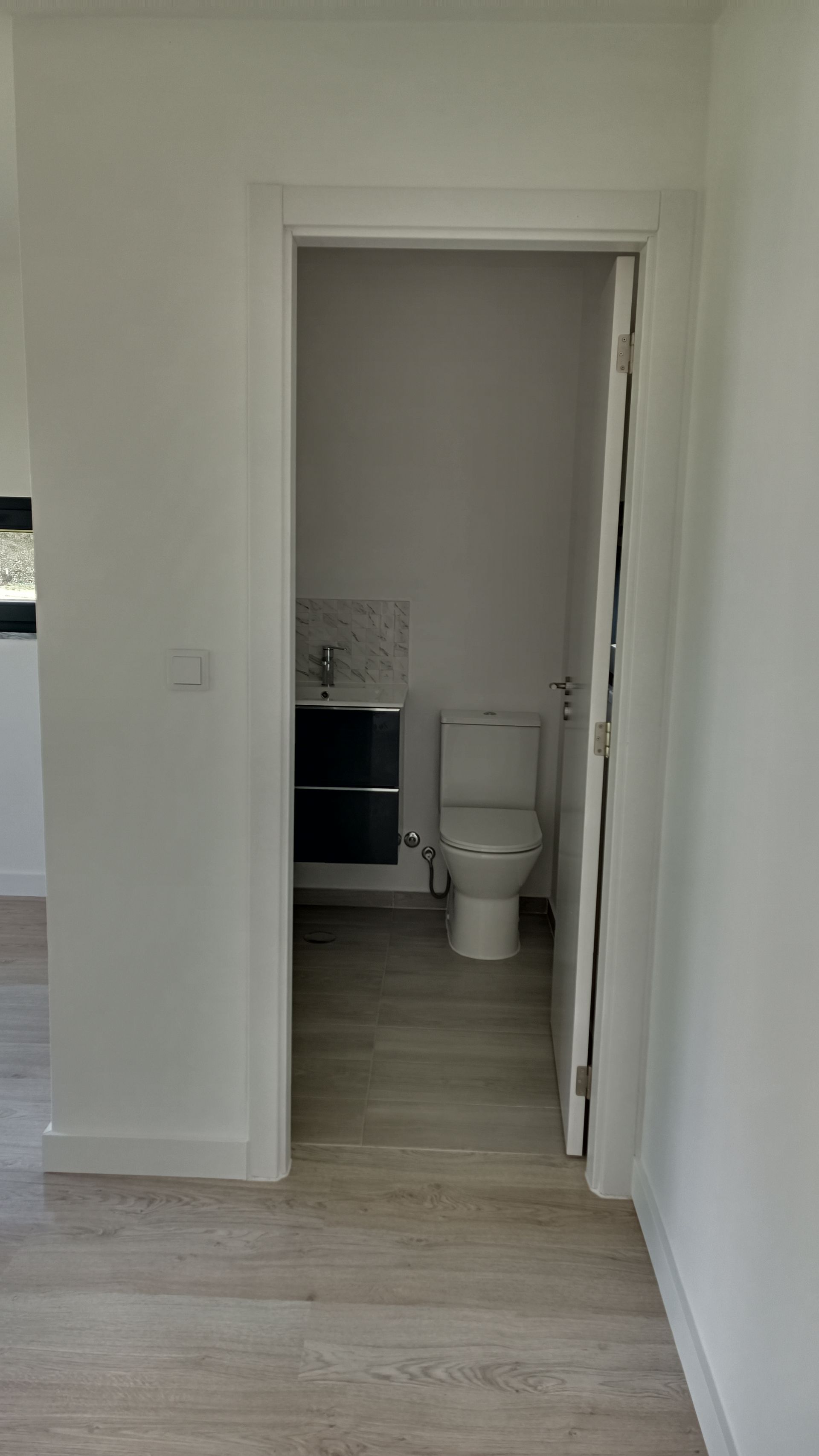 Vista de um banheiro pequeno. Paredes brancas, piso com aparência de madeira. Vaso sanitário e pia visíveis através da porta.