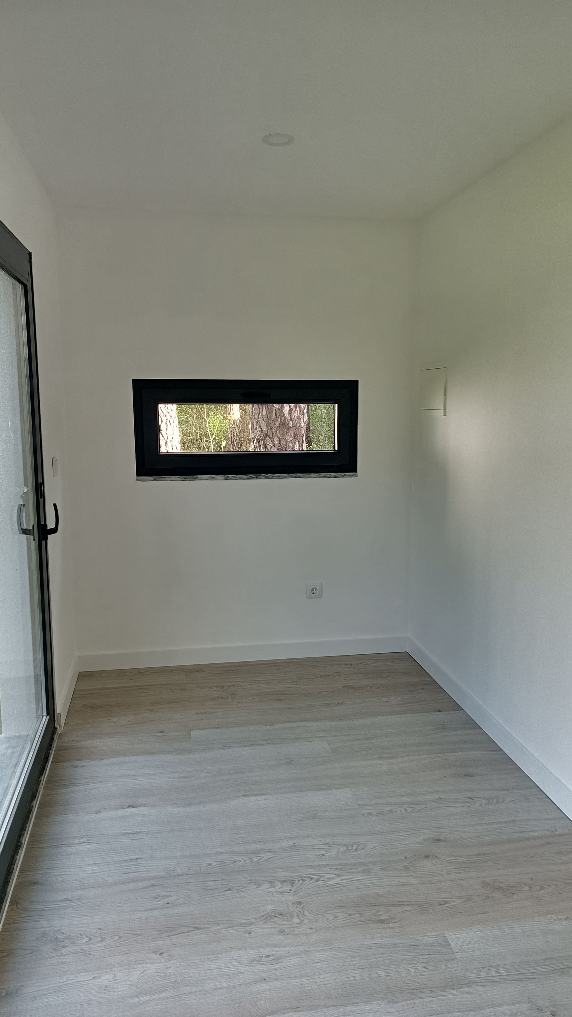 Quarto pequeno e vazio com paredes brancas, uma janela retangular estreita e uma porta de vidro.