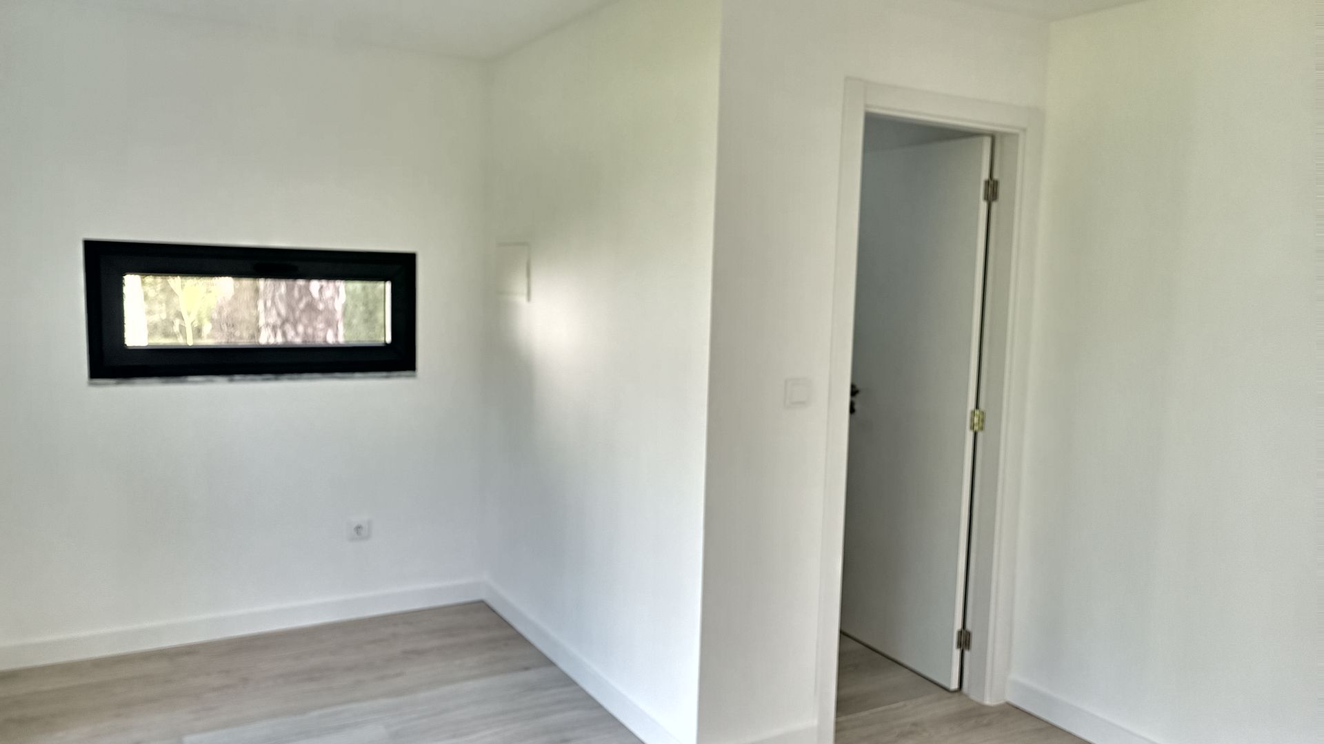 Quarto branco vazio com uma pequena janela preta e uma porta.