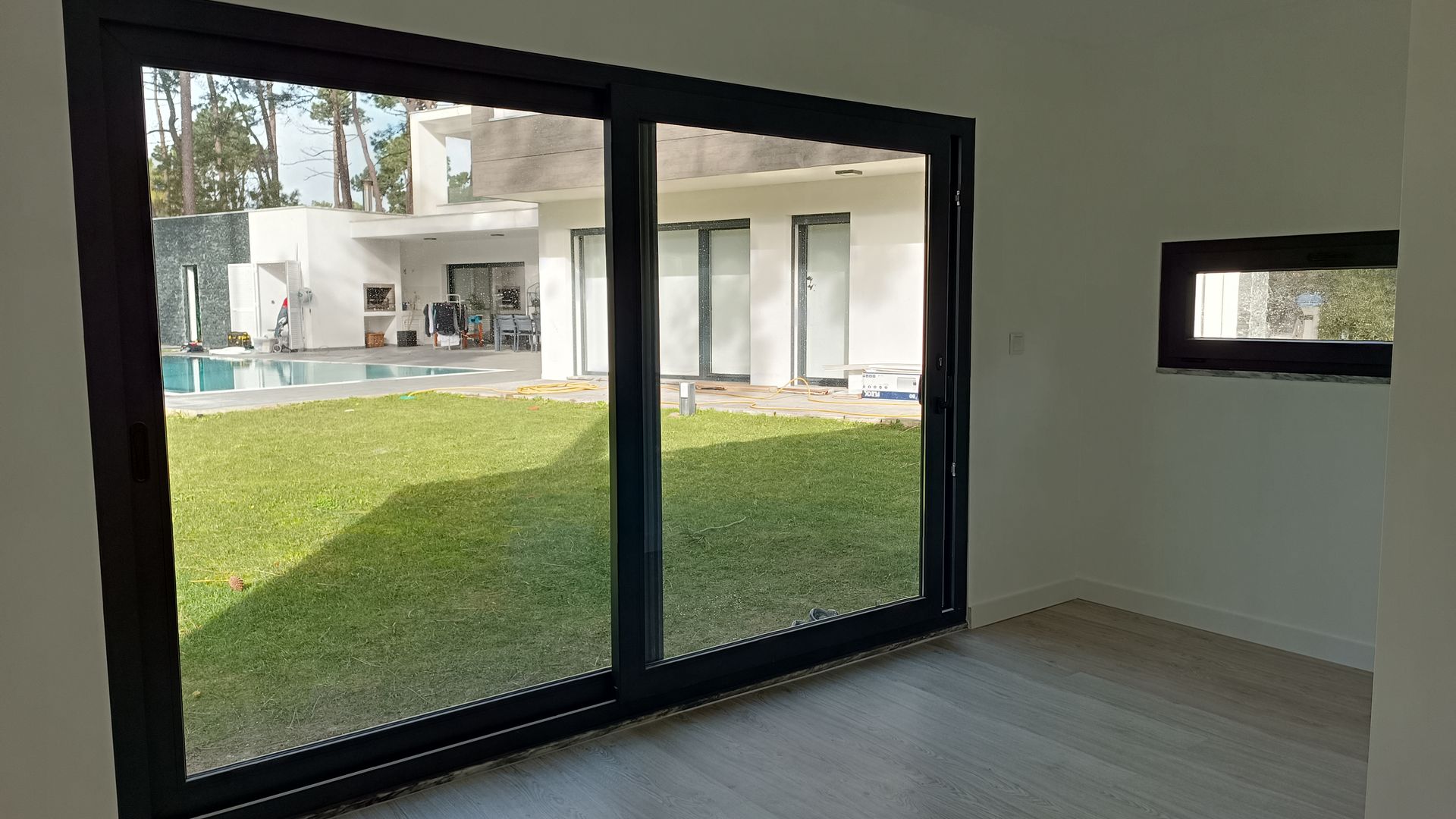 Vista interior de um cômodo com grandes portas de vidro emolduradas em preto, com vista para um gramado, piscina e uma casa moderna.
