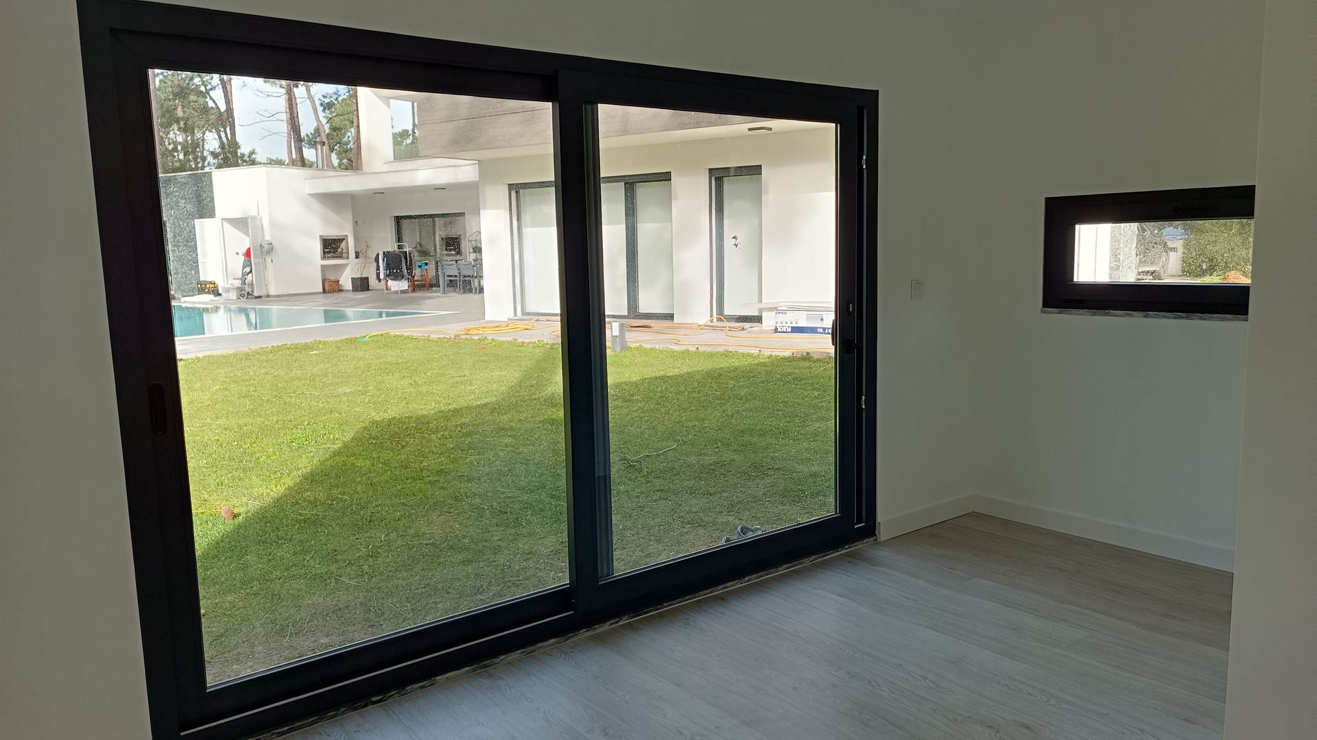 Vista interior de um quarto com grandes portas de vidro deslizantes com vista para um jardim verde com piscina.