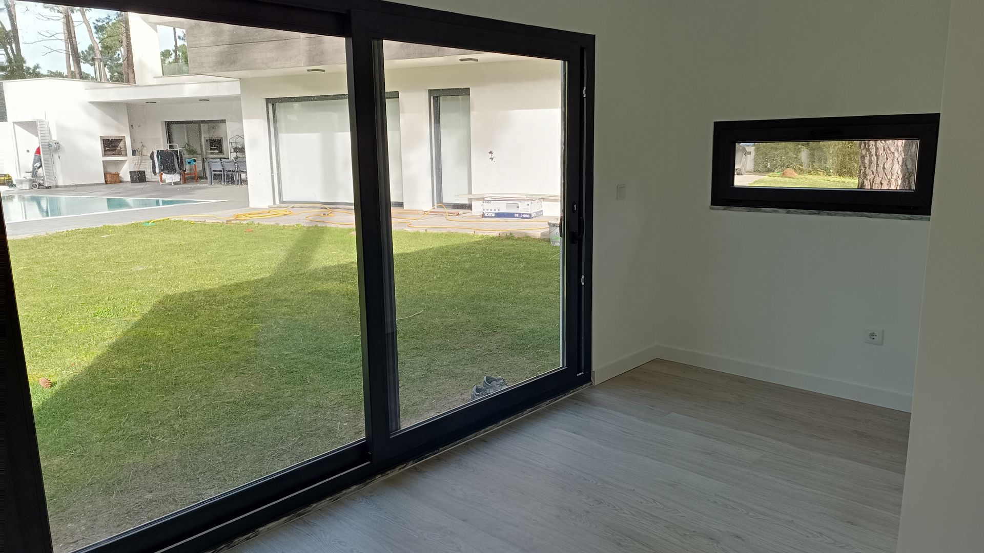 Interior de uma sala com grandes portas de vidro deslizantes com vista para um gramado verde, piscina e uma casa branca.