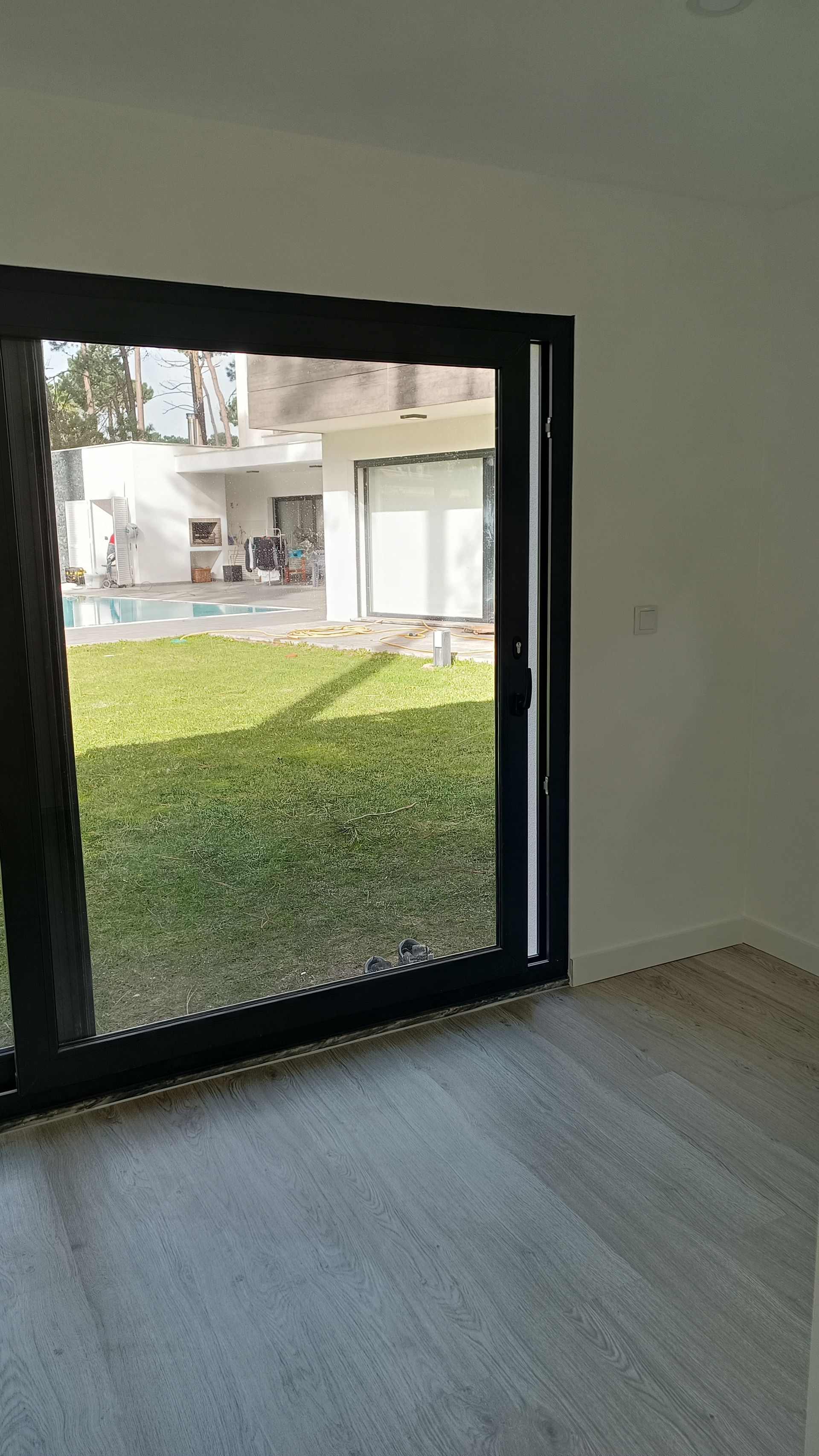 Vista interior de um cômodo com uma grande porta de vidro deslizante com moldura preta, com vista para um quintal gramado.