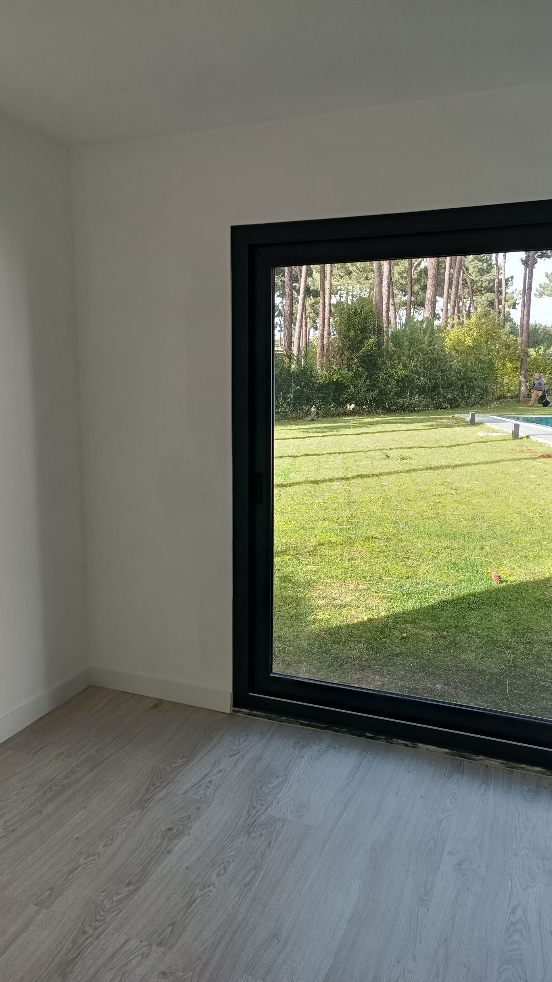 Quarto interior com piso de madeira clara, paredes brancas e grande janela com moldura preta com vista para um gramado verde.