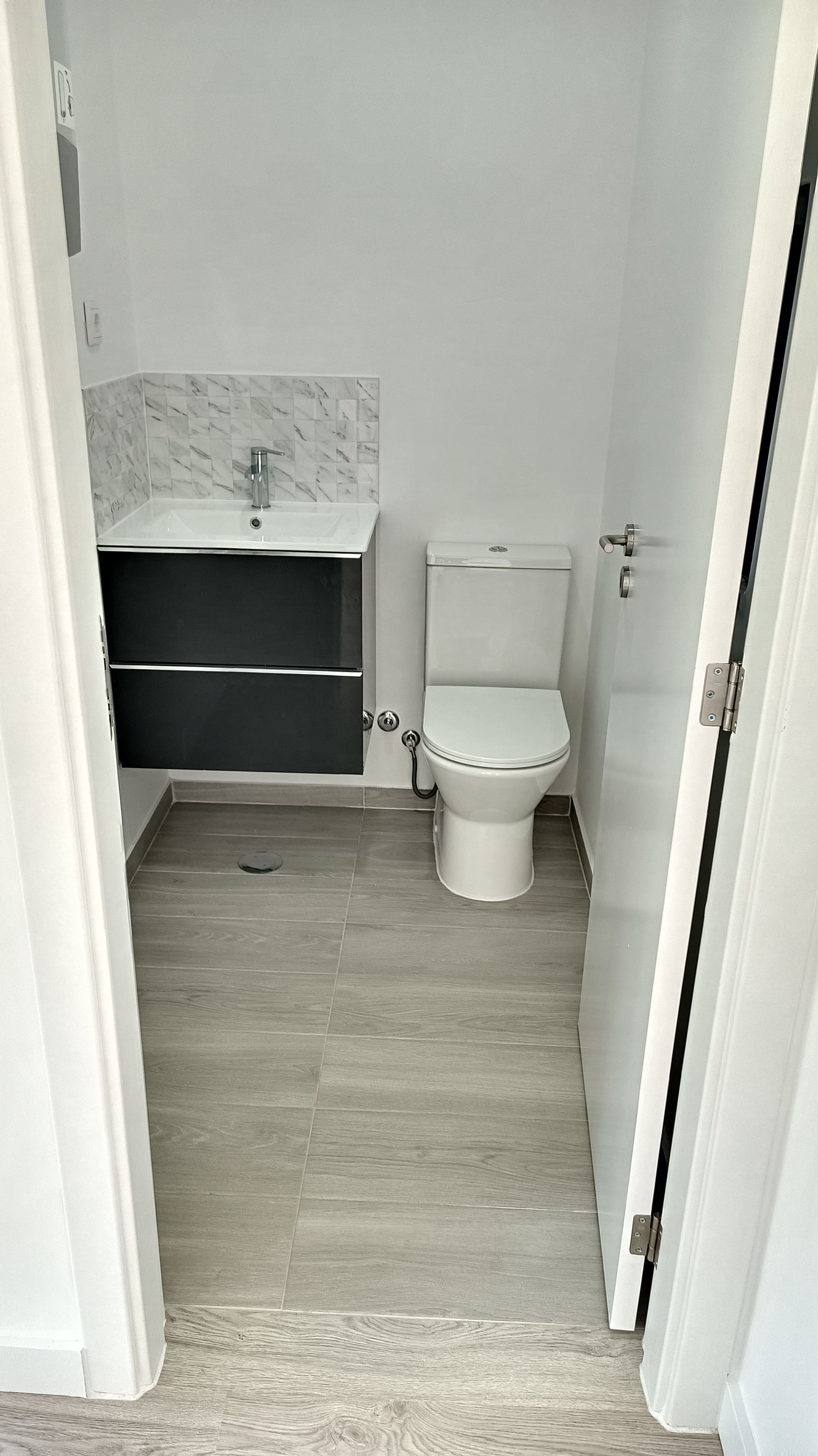 Banheiro estreito com piso de azulejo cinza, paredes brancas, vaso sanitário e pia com armário cinza.