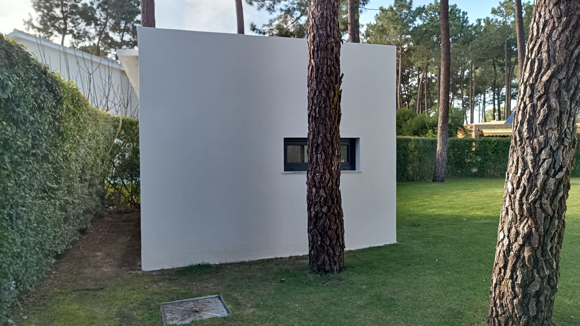 Prédio quadrado branco com uma pequena janela e uma árvore na frente, situado em um gramado verde.