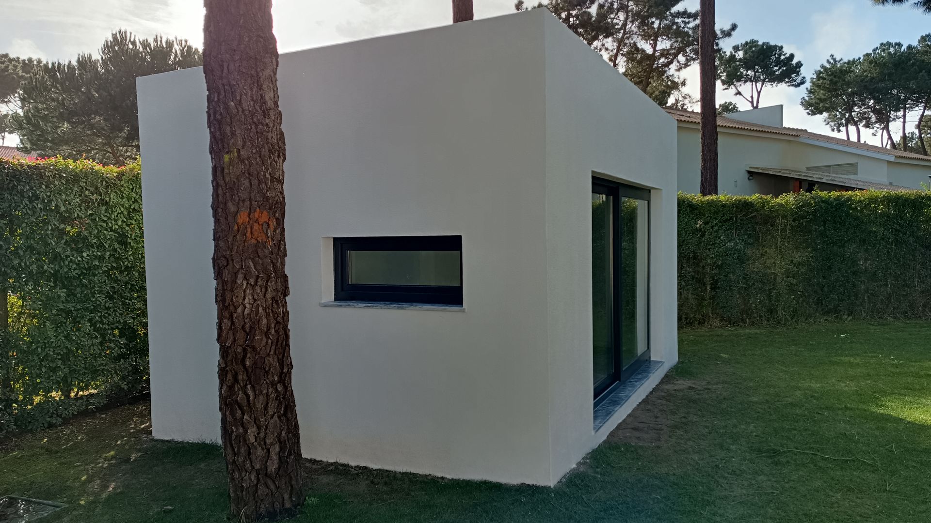 Pequeno edifício retangular branco com porta de vidro deslizante e janela pequena, ao lado de uma árvore e uma sebe.