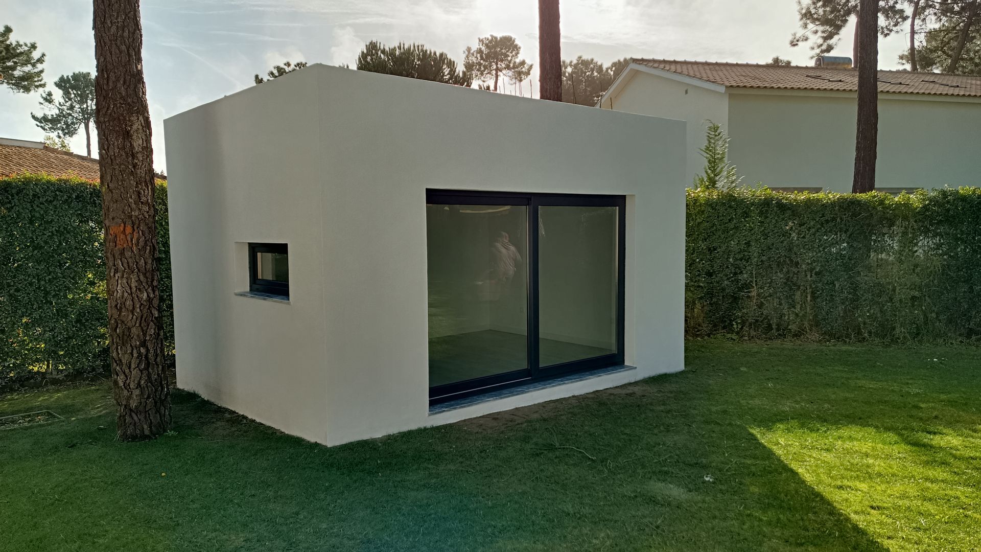 Casinha retangular branca com portas de vidro deslizantes emolduradas em preto e uma pequena janela, situada em um gramado verde.