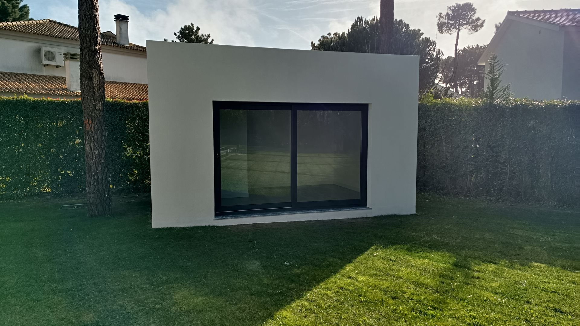 Prédio branco com portas de vidro deslizantes emolduradas em preto, cercado por grama verde e sebes.