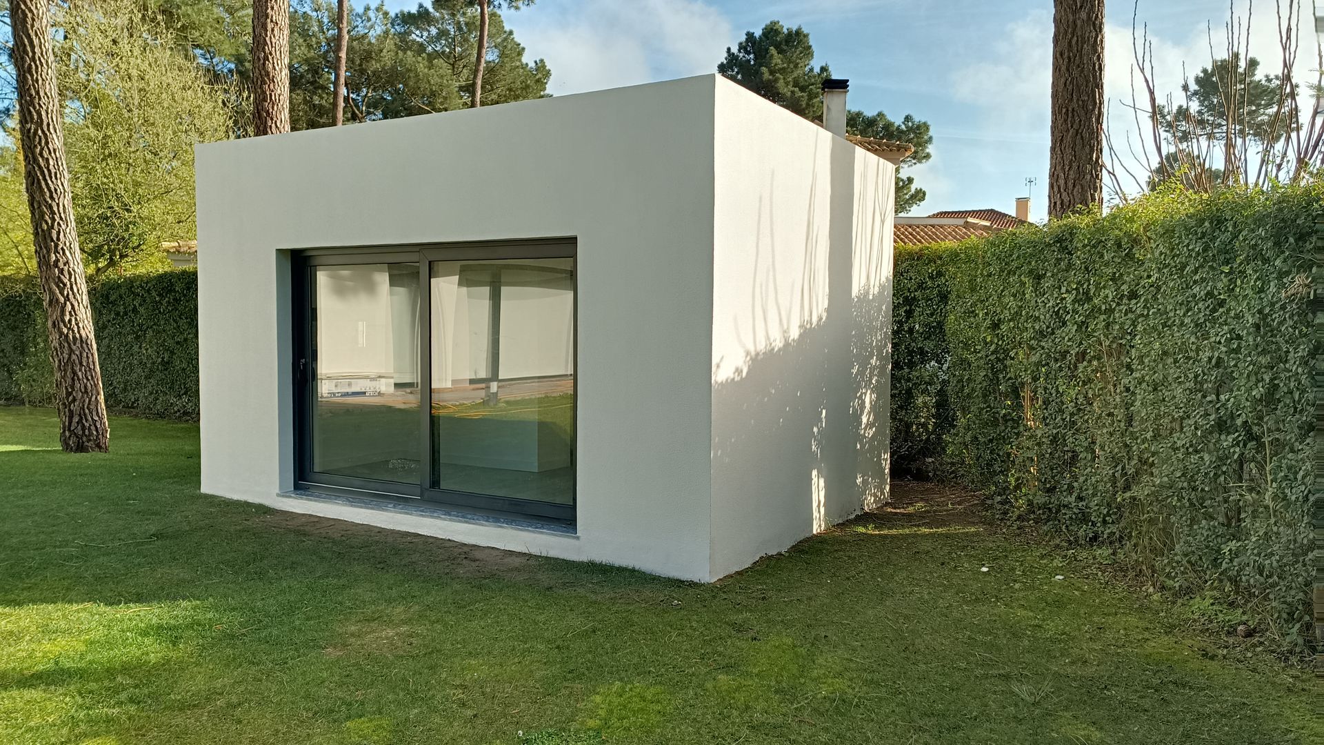 Edifício cinza em forma de cubo com grandes portas de vidro, rodeado por grama verde, sebes e árvores.