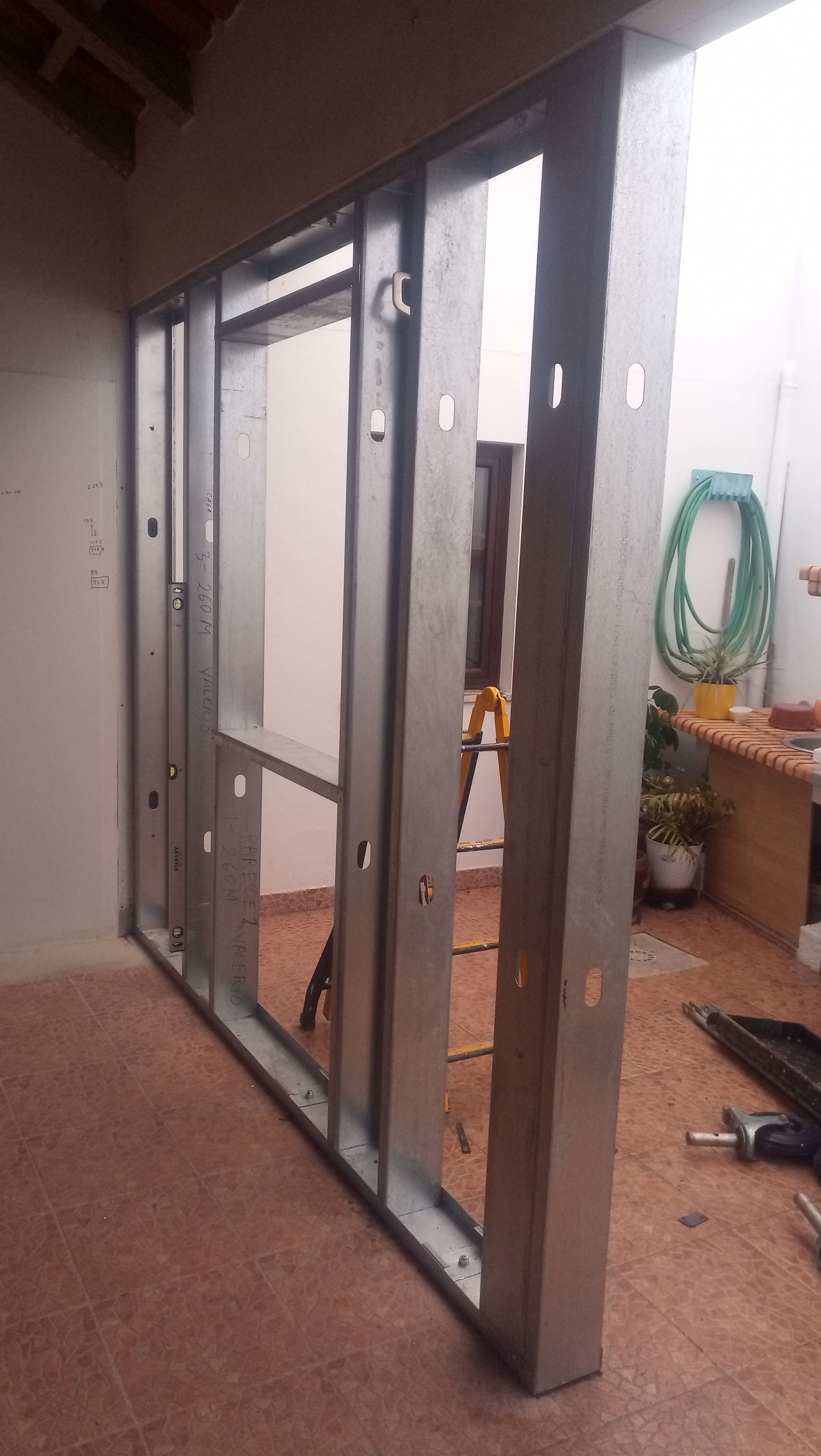 Estrutura metálica de uma parede em construção, com uma abertura para janela, em área externa sobre piso de azulejo.