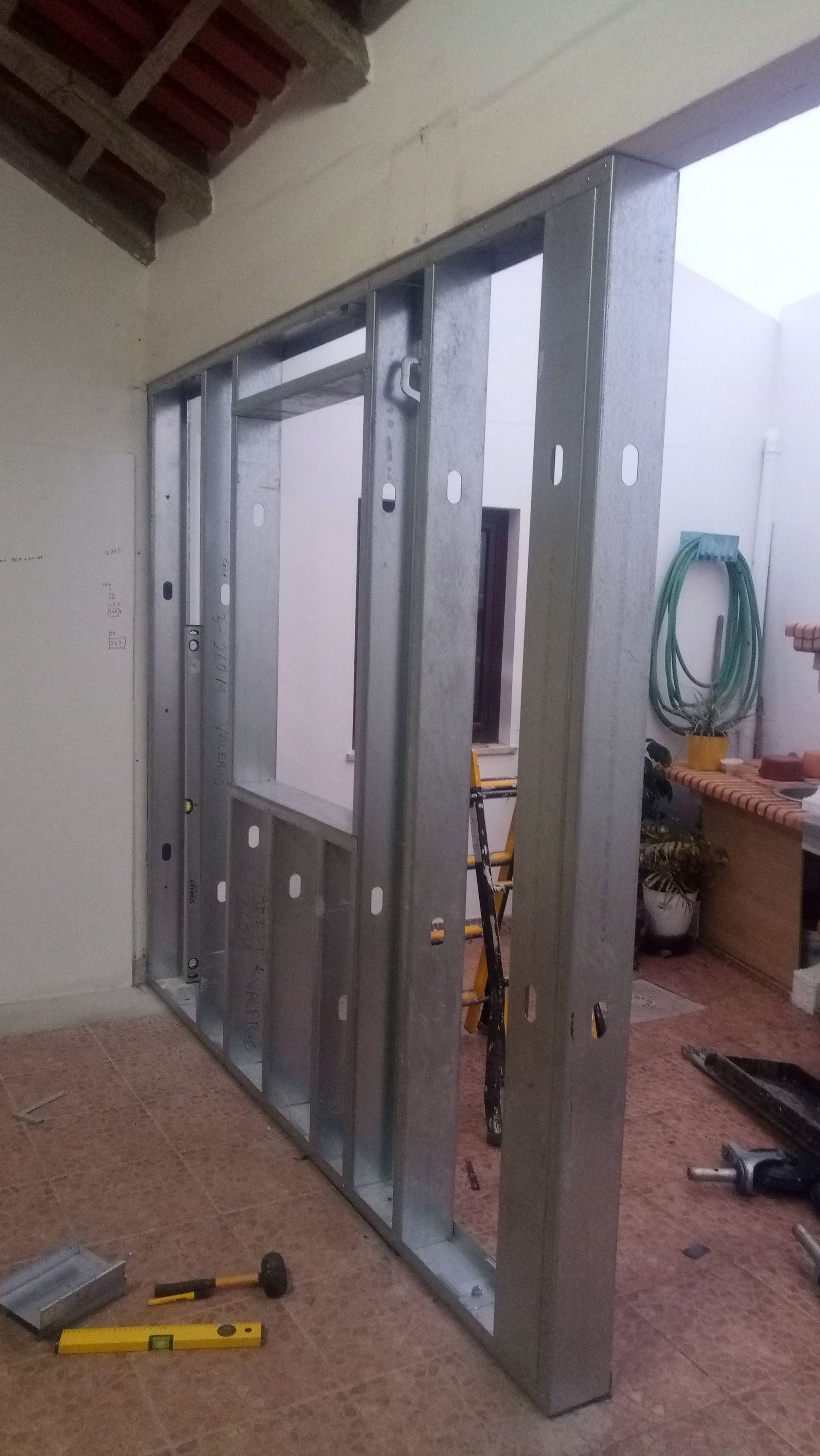 Estrutura metálica de uma parede em construção, com uma abertura para janela. Ferramentas espalhadas pelo chão em um ambiente interno.