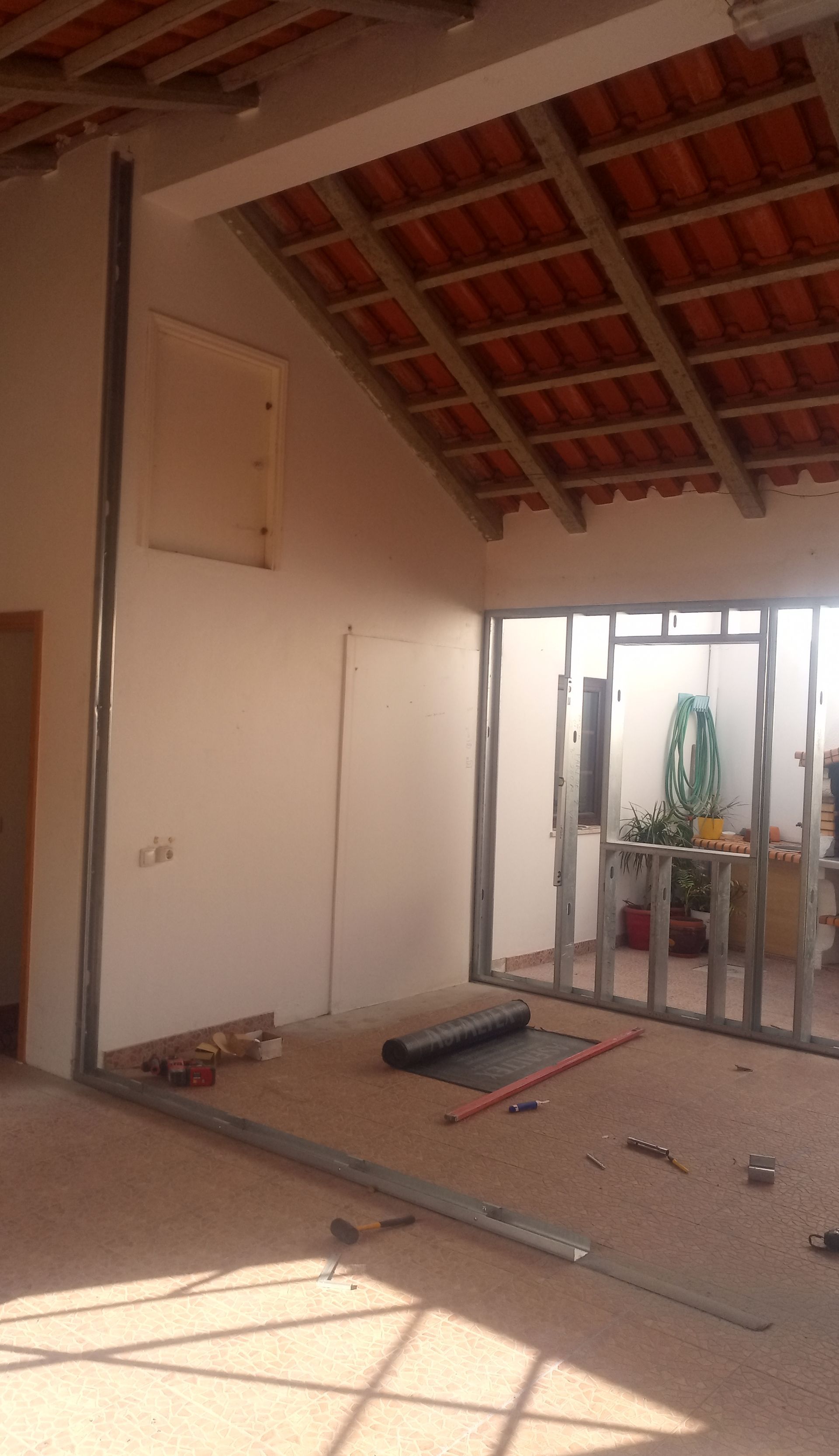 Vista interior de um cômodo em construção; perfis metálicos sustentam as paredes e uma abertura. Telhado de telha vermelha visível.