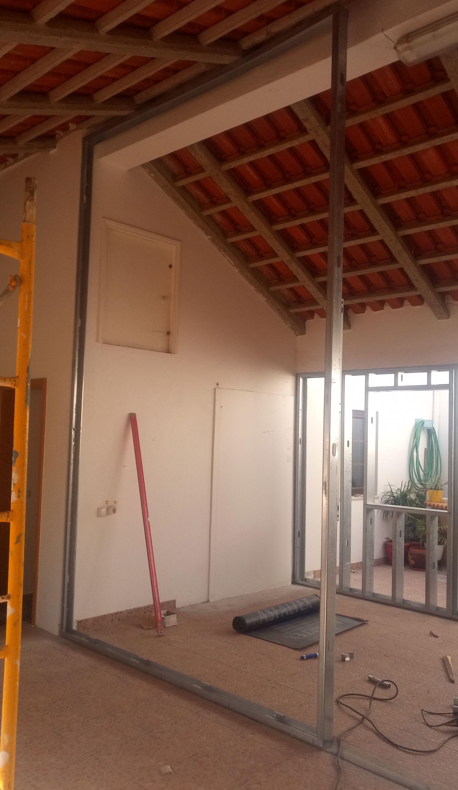 Interior em construção; estrutura metálica para as paredes, telhado de telha vermelha ao fundo, ferramentas no chão.